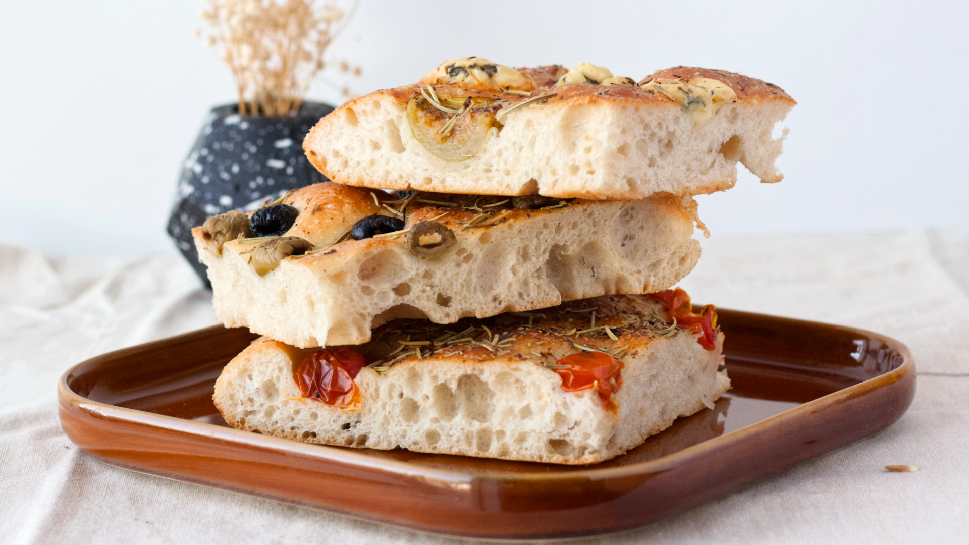 Zdjęcie przedstawia trzy kawałki focacci ułożone jeden na drugim. Za wierzchu focacci znajdują się dodatki: rozmaryn, pomidory, oliwki. Foccacie leżą na kwadratowym talerzu.