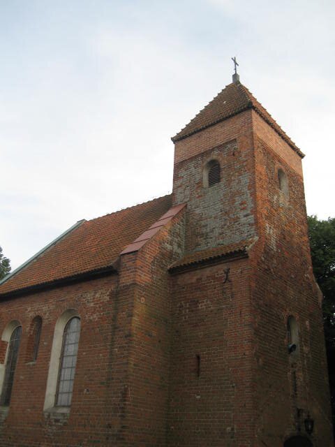 Zdjęcie przedstawia bok kościoła wykonanego z czerwonej cegły. Widoczna jest wieża umieszczona na przedzie, a także nawa główna ze skośnym dachem. Na jej ścianach znajdują się wysokie, podłużne okna. Na szczycie wieży znajduje się krzyż.