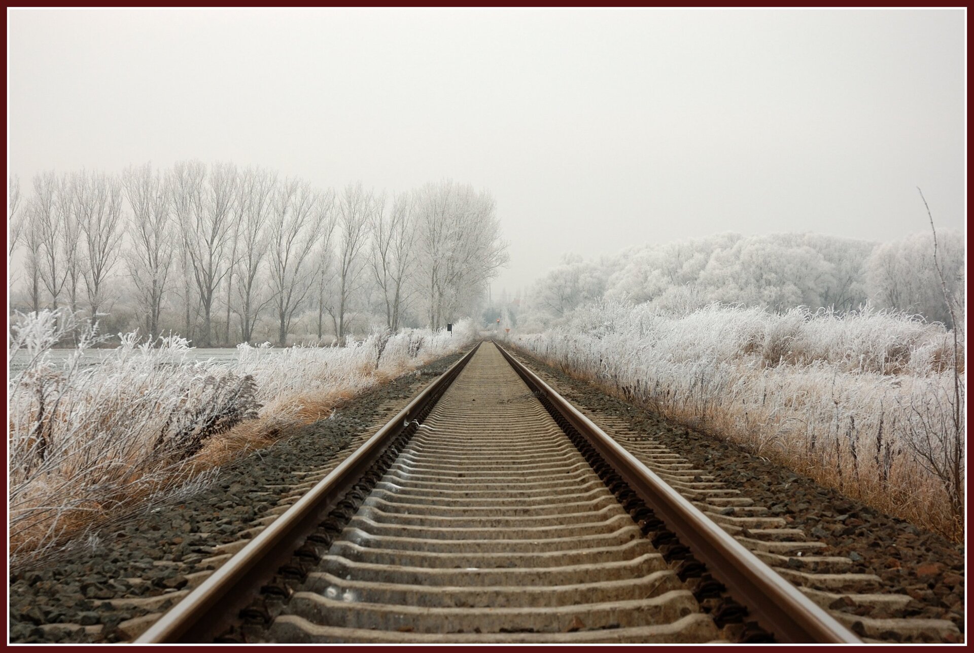 Ilustracja przedstawia tor kolejowy. Prowadzi, aż po horyzont, gdzie widać drzewa pokryte śniegiem. Prowadzi on przez łąkę i las. Pora roku przedstawiona na zdjęciu to zima. Niebo pokryte jest jasnymi białymi chmurami.