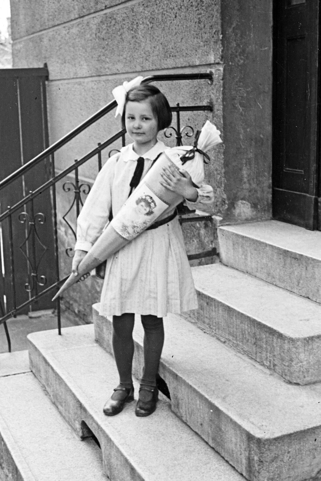 Zdjęcie jest czarno‑białe. Przedstawia uśmiechniętą, elegancko ubraną dziewczynkę, na głowie ma kokardę. W dłoniach trzyma duży rożek wypełniony słodyczami, starannie opakowany.