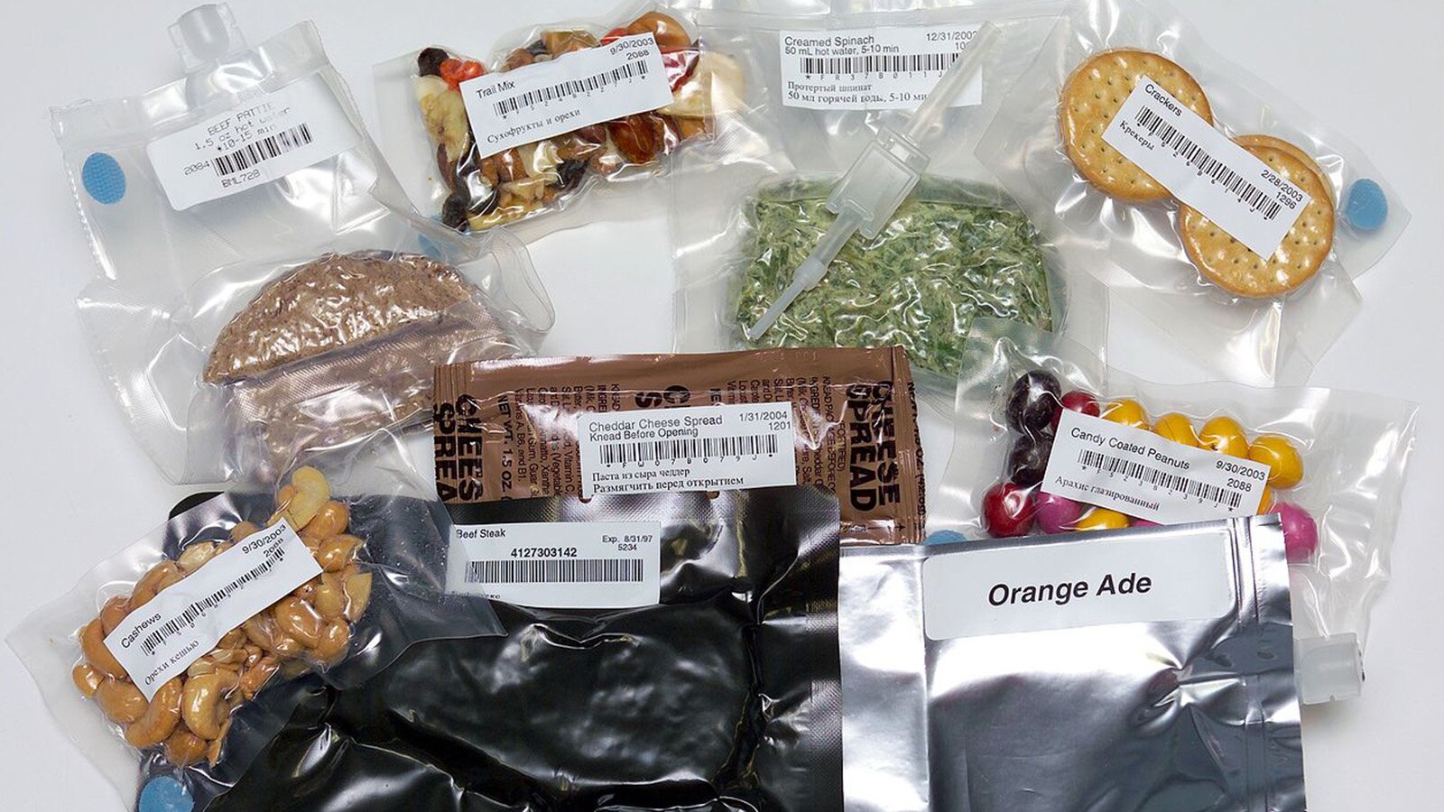 Zdjęcie przedstawia różne rodzaje jedzenia, zapakowane próżniowo w foliowe, przeźroczyste woreczki lub woreczki ze srebrnej folii. Każdy woreczek ma białą etykietę z napisem lub napisem i kodem kreskowy. Napisy są w języku angielskim oraz rosyjskim. Na etykietach widoczne są napisy takie jak:
BEEF PATTIE, TRIAL MIX, CREAMED SPINACH, CRACKERS, CHEDDAR CHEESE SPREAD, CANDY COATED PEANUTS, ORANGE ADE, BEEF STEAK, CASHEWS.