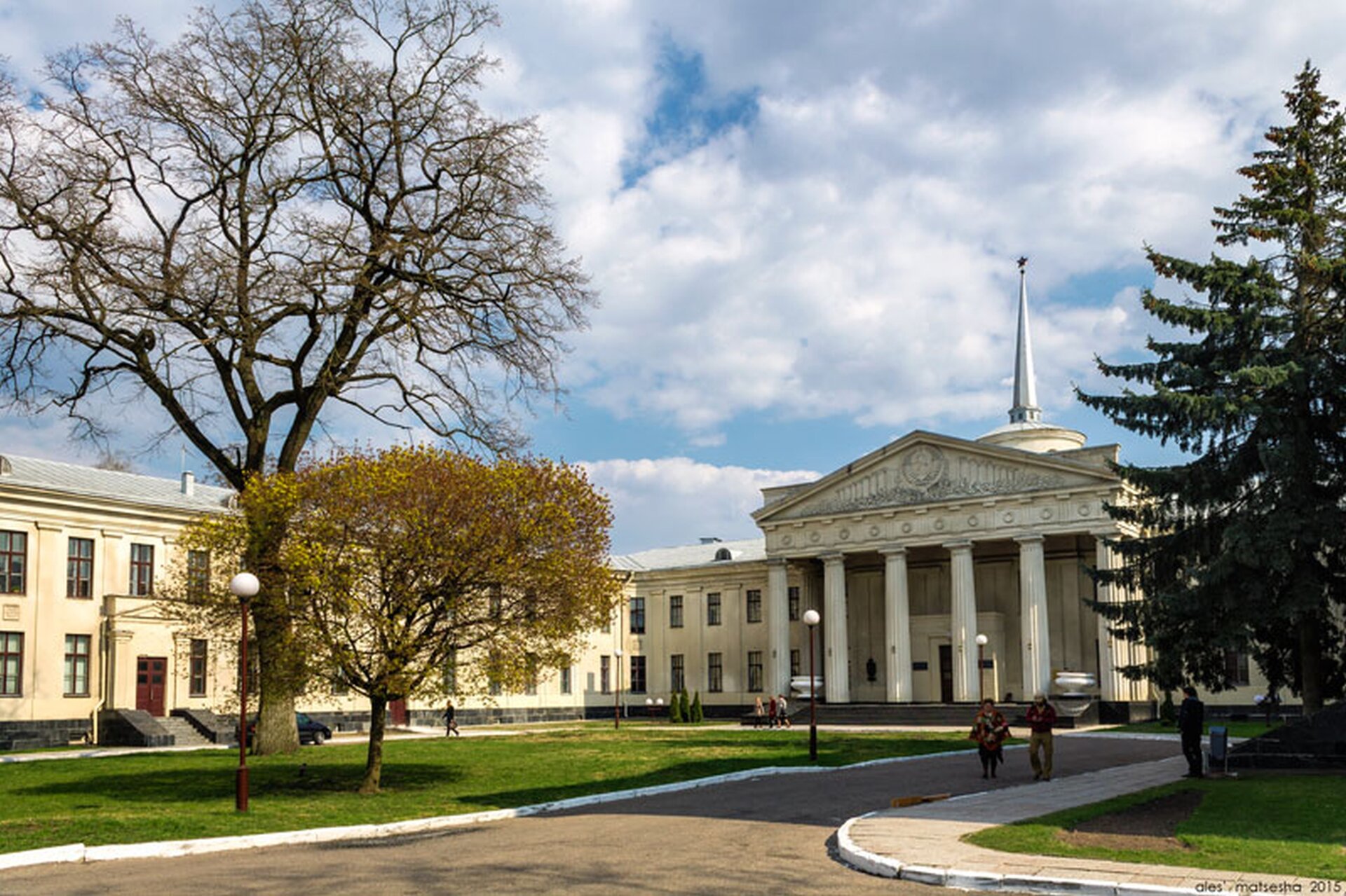 Zdjęcie dziedzińca porośniętego trawą, dookoła droga i chodnik. Przed dziedzińcem okazały pałac z kolumnami. 