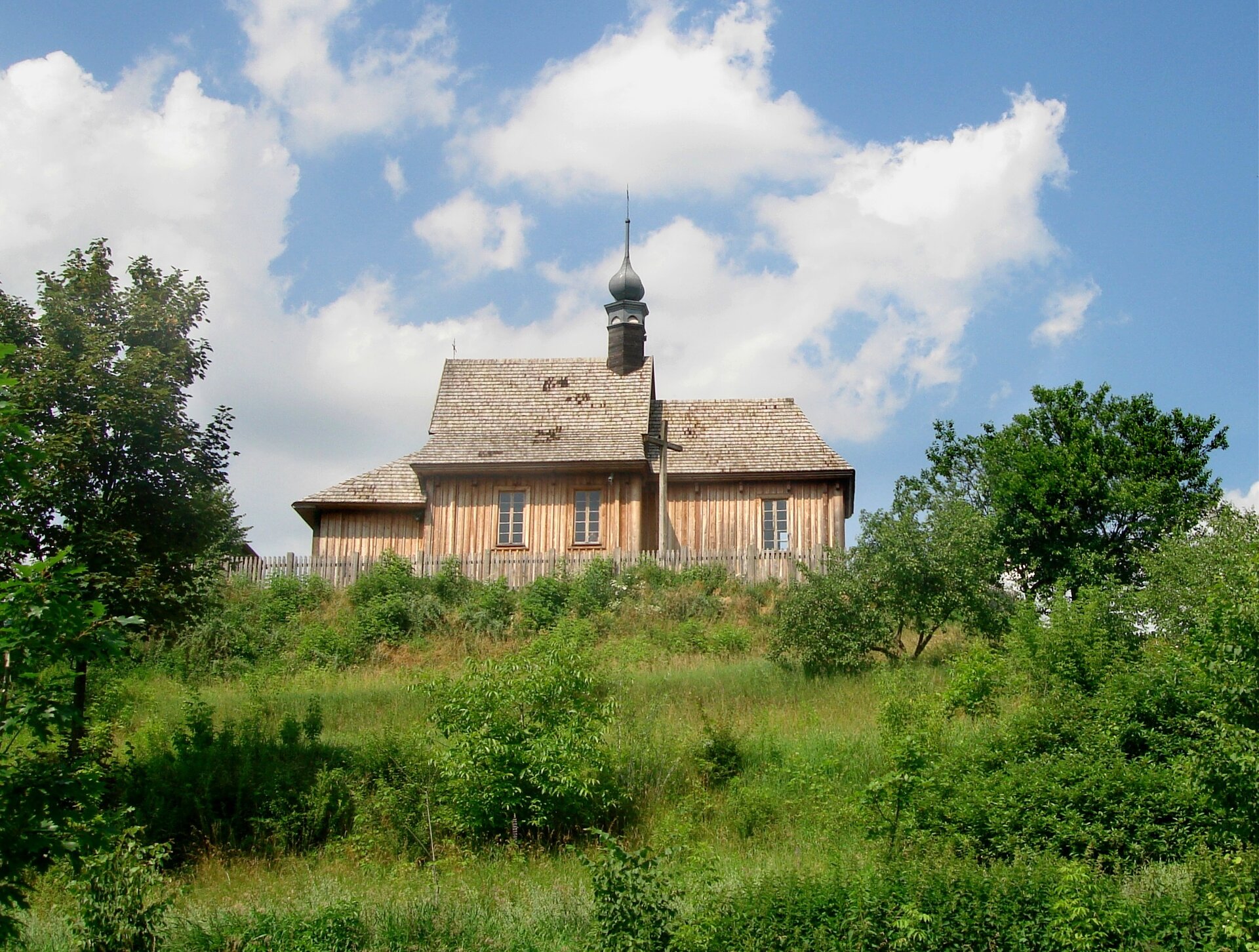 Zdjęcie przedstawia niewielki budynek zbudowany na wzniesieniu. Ma jedną wieżę z krzyżem. Dookoła zielona roślinność.
