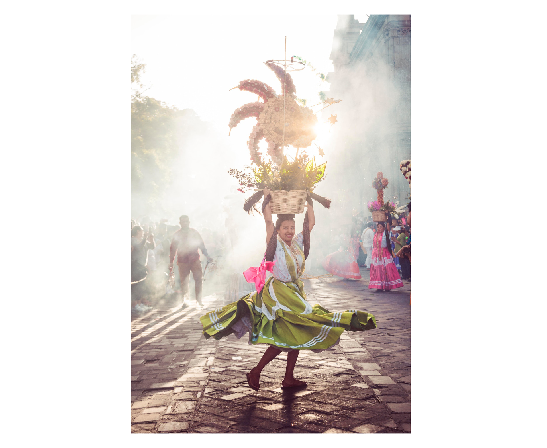 Zdjęcie przedstawia ulicę, na której odbywa się festyn. Na pierwszym planie ukazana jest młoda, uśmiechnięta kobieta w jasnej bluzce i szerokiej, oliwkowej spódnicy. Kobieta ma długi warkocz, w który wpleciona jest różowa wstążka. Kobieta trzyma na głowie duży, wiklinowy kosz wypełniony różnorodnymi, kolorowymi polnymi kwiatami, a nad nim ułożone z kwiatów słońce. W tle widoczne są kolejne kobiety trzymające na głowach kosze z kwiatami. Ulicę spowija jasny dym. Wokół kobiet z koszami stoi tłum ludzi.