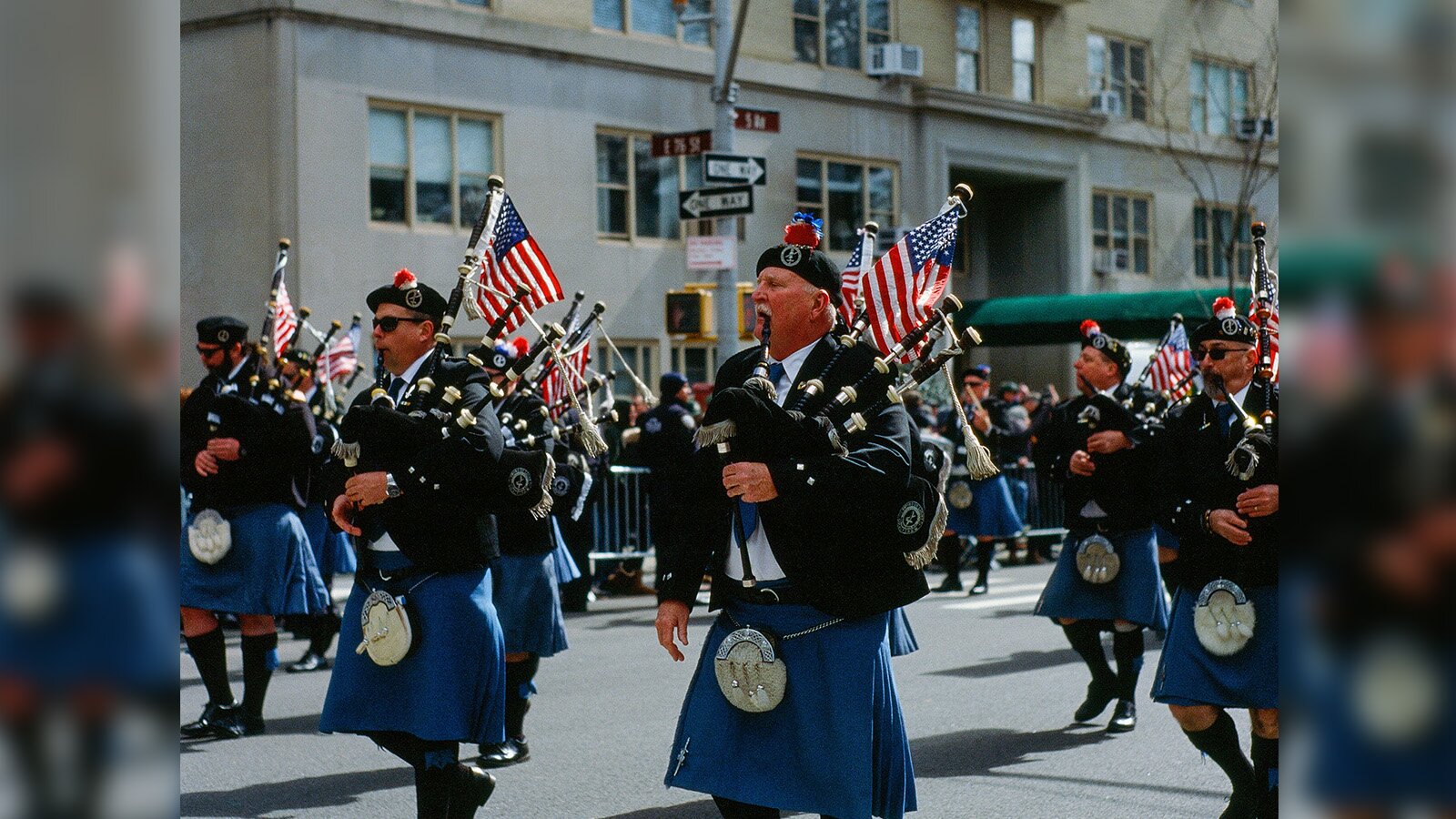 Zdjęcie przedstawia paradę maszerującą ulicą w lewo. Widoczni są mężczyźni idący w formacji, ubrani w ciemnoniebieskie kitle, białe koszule, granatowe marynarki oraz czarne berety z pomponikami. Każdy z mężczyzn trzyma dudy i gra na nich. Do pionowego elementu każdego instrumentu zamocowana  jest flaga Stanów Zjednoczonych.