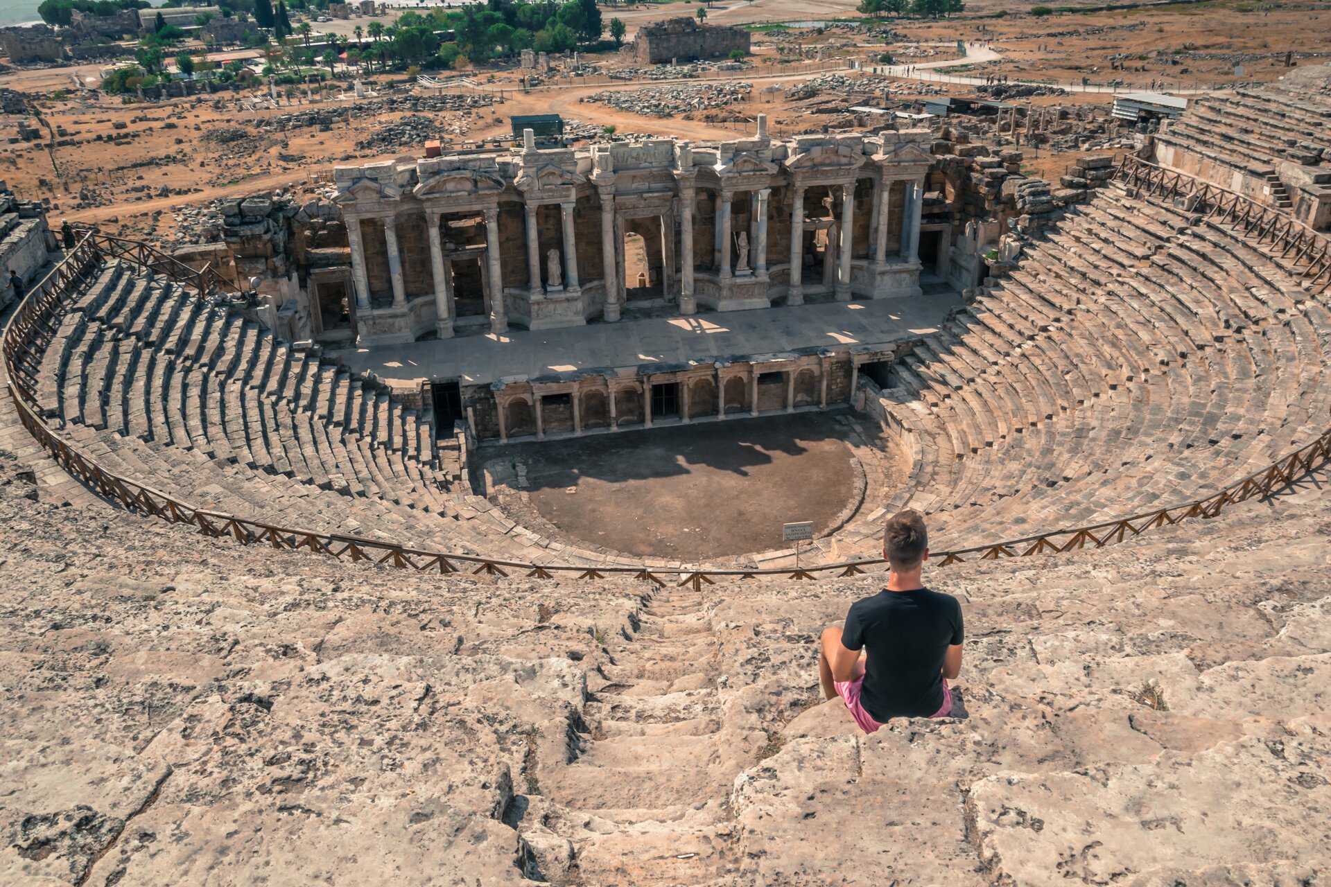 Fotografia przedstawia widok na pozostałości po starożytnym teatrze. Scena umieszczona jest na półkolu wewnątrz widowni, na której siedzi mężczyzna ubrany w czarną koszulkę oraz różowe spodnie. Widownia jest podzielona na sekcje za pomocą ogrodzenia. Za sceną znajdują się ruiny budynku, który służył jako zaplecze i garderoba.  