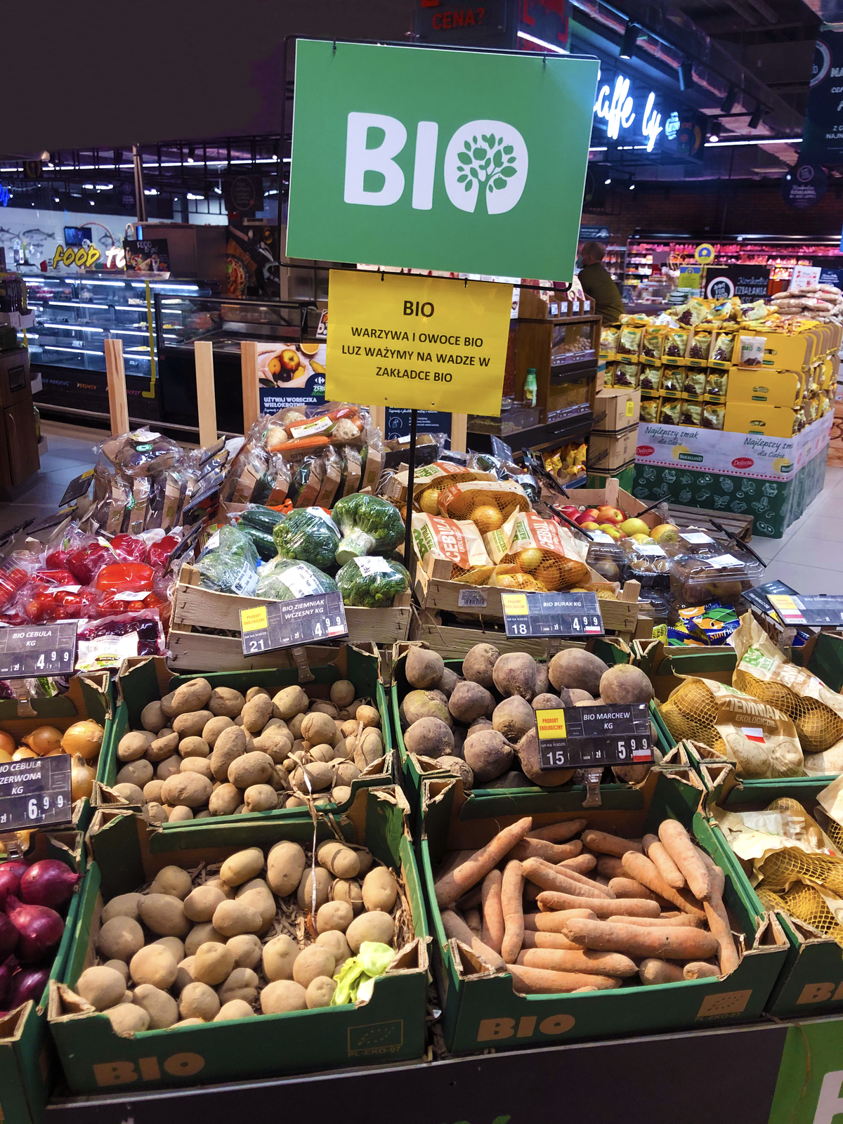 Na ilustracji widnieje stoisko z żywnością BIO w supermarkecie. Są to np. marchewki, ziemniaki, buraki, kalafiory, brokuły, papryki, cebule. 