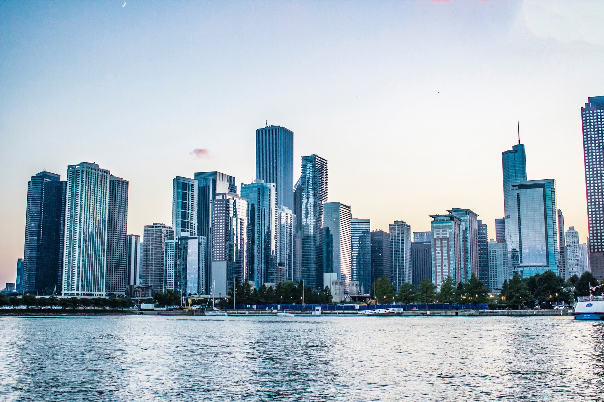 Na ilustracji przedstawiono panoramę miasta, za rzeką. Widoczne wysokie biurowce oraz drapacze chmur.