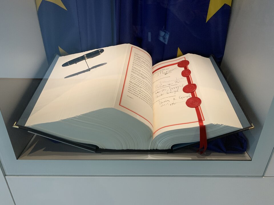 Zdjęcie przedstawia grubą księgę, która leży otwarta w szklanej gablocie. Widoczny jest tekst, odręczne podpisy oraz czerwone pieczęcie. Na stojaku znajduje się pióro.