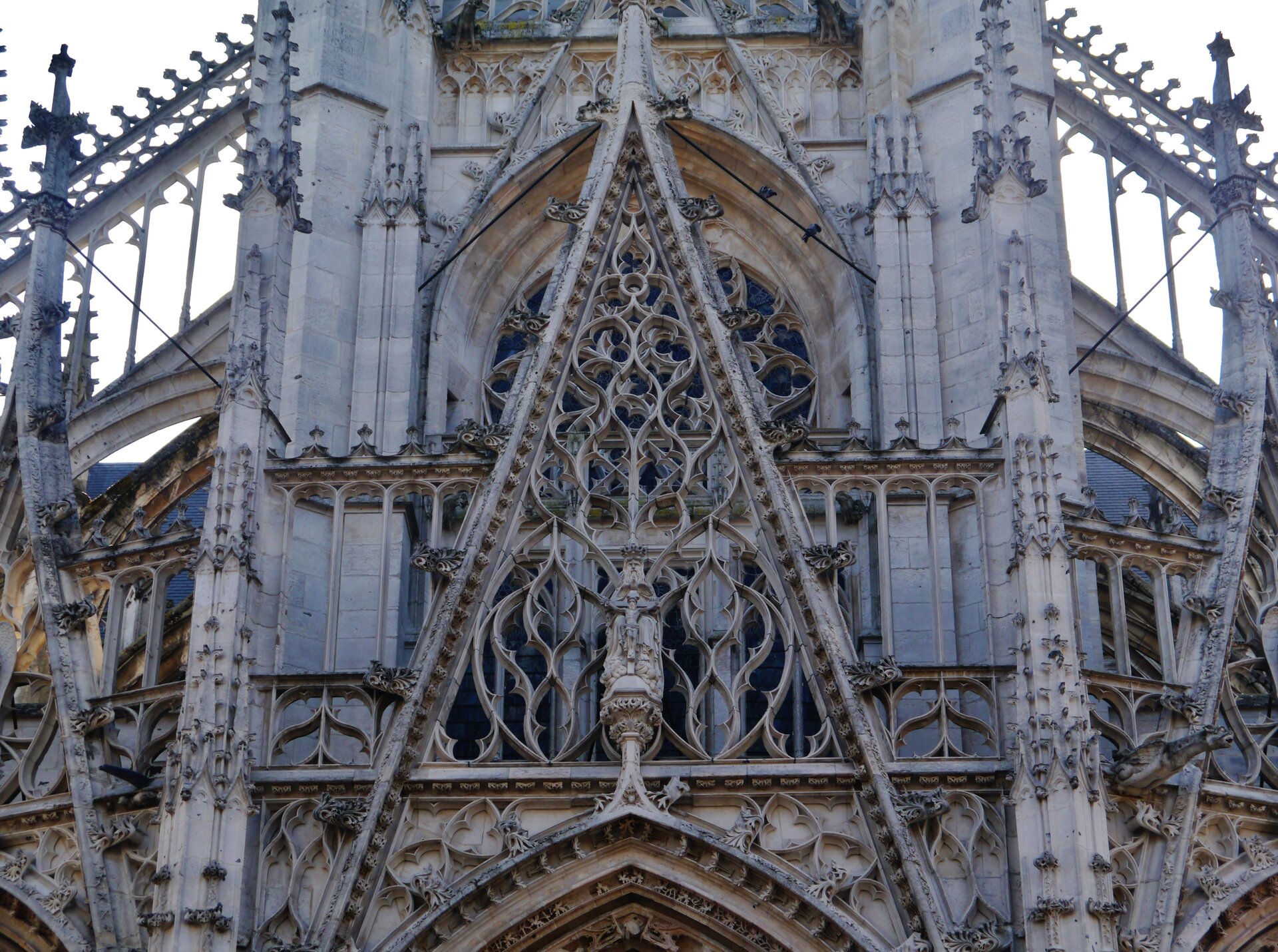 Fotografia przedstawia detal budynku - wimpergę - element architektoniczny o kształcie trójkąta, który zdobi fasadę budynku. Element ten został umieszczony nad oknem witrażowym - posiada ażurową konstrukcję, która swoim kształtem i wzorem przypomina łodygę z cierniami. W wewnętrznych krawędziach umieszczono zdobienia, kształtem przypominające stożki oraz ornamenty liściaste. Wewnątrz ażurowej konstrukcji - Jezus wiszący na krzyżu oraz Bóg Ojciec czuwający nad ciałem swojego syna.