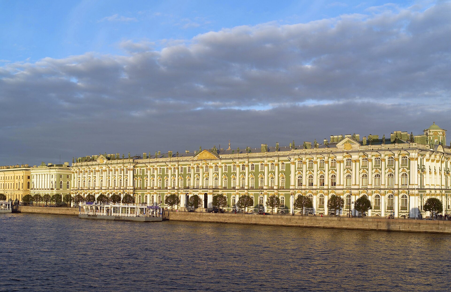 Na zdjęciu znajduje się Pałac Zimowy, jeden z budynków muzeum w Petersburgu. Pałac, zbudowany w stylu barakowym, jest położony na brzegu Newy. To prostokątny gmach z  prostokątnymi oknami, zakończonymi łukami i naprzemiennie występującymi wypukłymi i wklęsłymi elementami. Pomiędzy oknami są pilastry. Dwa pilastry, zwieńczone tympanonem z frontonem, są ozdobione herbami. Dach zwieńczony jest balustradową attyką.