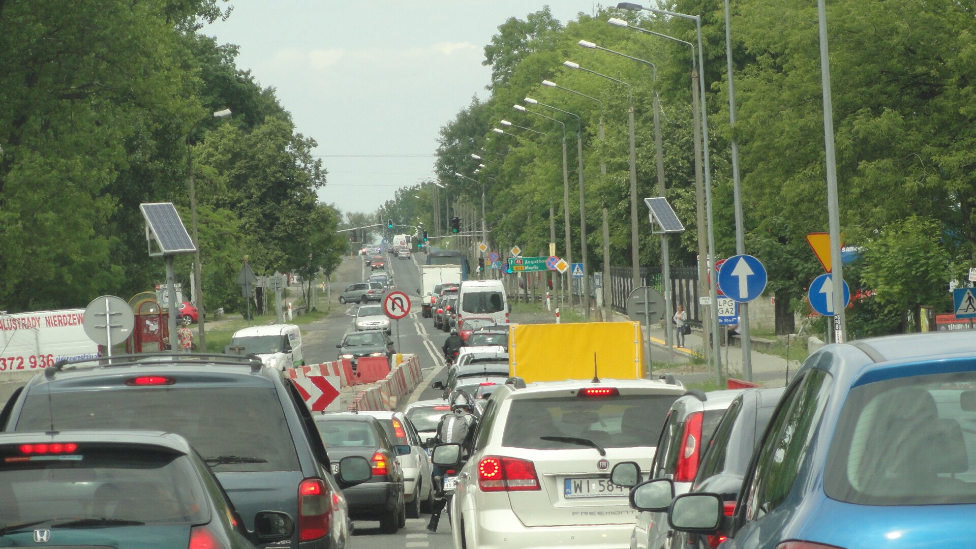 Zdjęcie przedstawia korek na drodze spowodowany robotami drogowymi. Przez środek zdjęcia przebiega ulica, która jest zapełniona przez samochody. W centrum znajdują się biało‑czerwone barierki, które odgradzają część pasa ruchu, przez co dwa po prawej stronie zwężają się do jednego. Po obu stronach ulicy rosną drzewa.