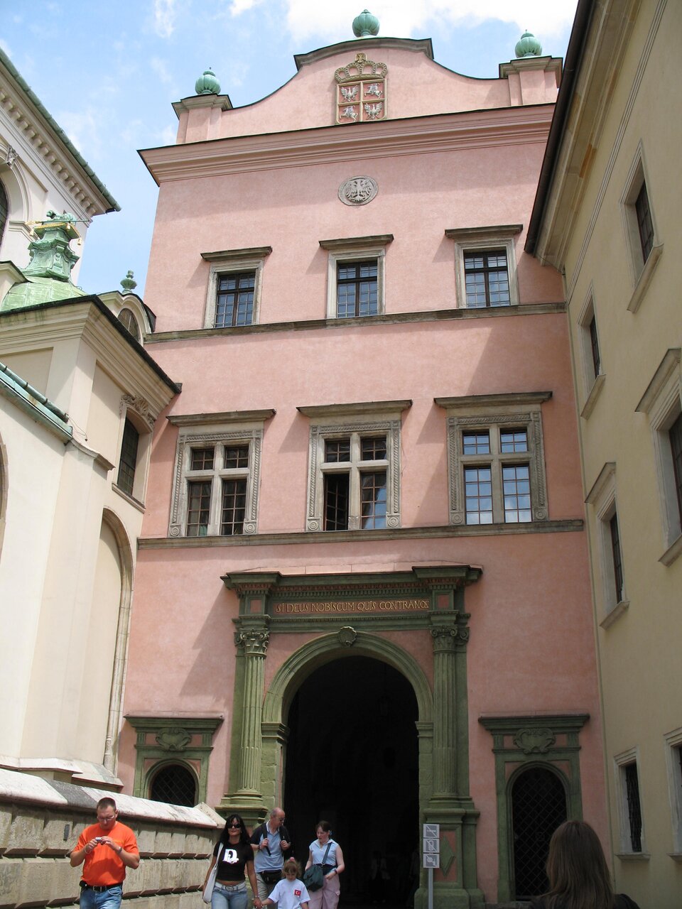 Ilustracja przedstawia bramę wjazdową do zamku królewskiego na Wawelu. Budynek jest dwupiętrowy z trzema oknami na każdym piętrze, zwieńczony attyką i wjazdem zwieńczonym półokrągłym łukiem. Znajduje się pomiędzy zwartą zabudową.