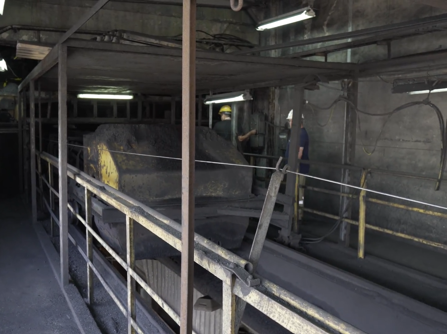 Na zdjęciu widoczna jest nasypywarka. Nasypywarka znajduje się wewnątrz budynku. Nasypywarka ma kształt prostopadłościanu o zaokrąglonych wierzchołkach. Element znajduje się nad kanałem. Po prawej stronie nasypywarki widoczni są dwaj pracownicy ubrani w stroje robocze.