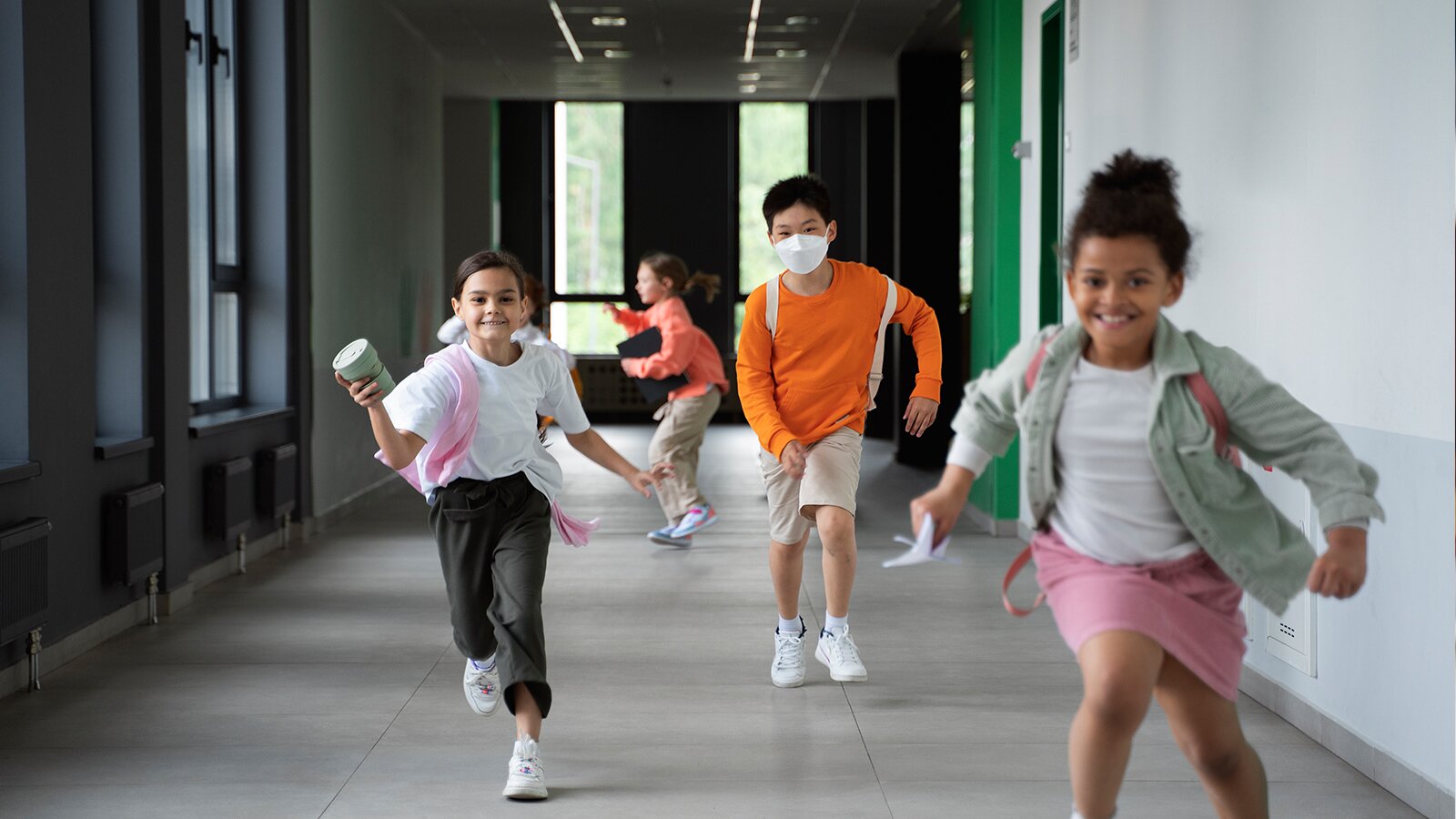 Zdjęcie przedstawia czworo uśmiechniętych dzieci w wieku około dziesięciu lat, biegnących po korytarzu. Wszyscy maja na sobie białe adidasy i plecaki.