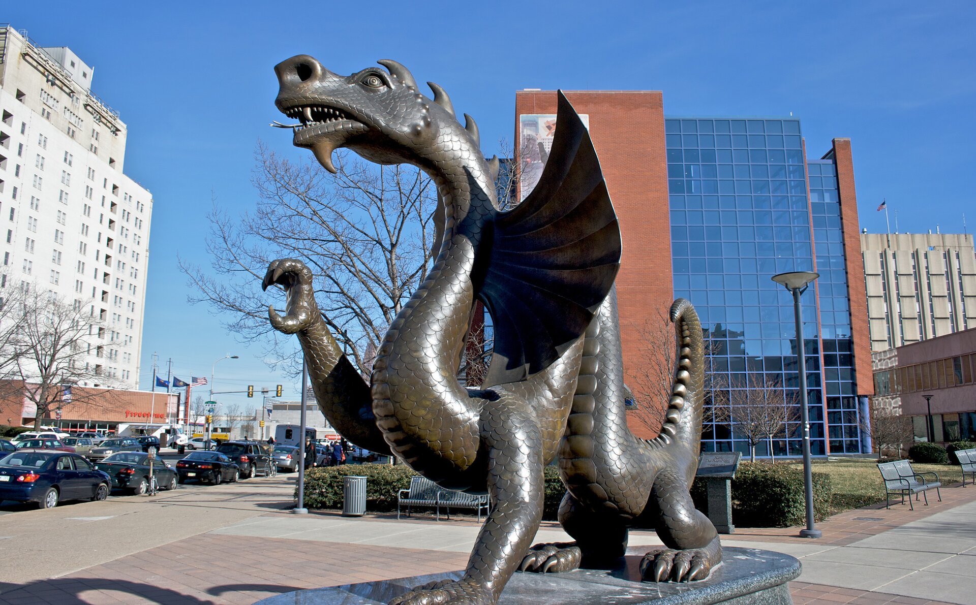 Kliknij, aby powiększyć. Zdjęcie przedstawia Mario Wspaniałego, maskotkę Uniwersytetu Drexel w Stanach Zjednoczonych. Rzeźba ze srebrzystego metalu. Smok przypomina węża na czterech lapach. jedną łapę ma uniesioną z wyciągniętymi pazurami. Po bokach szyi skrzydła. Psyk półotwarty, widoczne liczne ostre zęby. Na ciele szereg wypustek. W tle smoka widać różne budynki - budynek po lewej stronie jest biały, budynek po prawej stronie jest w połowie ceglany, a w połowie przeszklony. Widać latarnie uliczne i zaparkowane wzdłuż ulicy samochody.