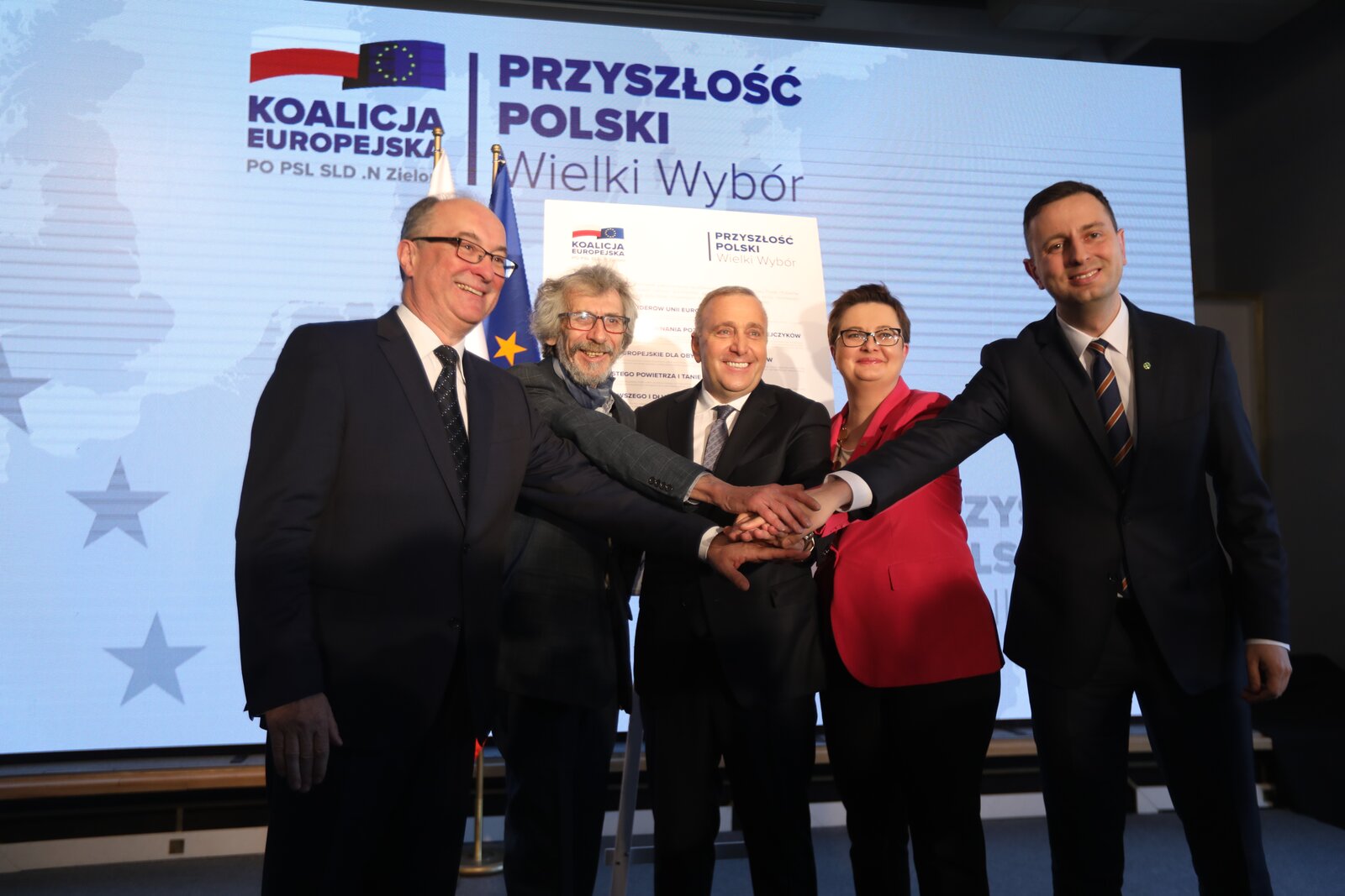 Zdjęcie przedstawia pięcioro polityków. Czterech mężczyzn (Włodzimierza Czarzastego, Dariusza Szweda, Grzegorza Schetynę, Władysława Kosiniaka-Kamysza) i jedną kobietę (Katarzynę Lubnauer). Wszyscy stoją obok siebie i trzymają dłoń na dłoni.