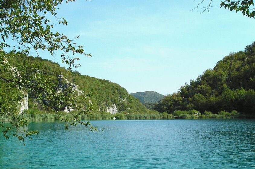 Zdjęcie przedstawia krajobraz Alp Dynarskich. W Alpach Dynarskich w Chorwacji panuje chłodniejszy i bardziej wilgotny klimat, dzięki czemu można tam rosnąć lasy liściaste. The Dinaric Alps in Croatia have a cooler and more humid climate which makes it possible for broadleaf forests to grow there. Na pierwszy planie widoczny jest zbiornik wodny. Z brzegów jeziora wyrastają niezbyt wysokie skaliste wzgórza porośnięte częściowo roślinnością. 