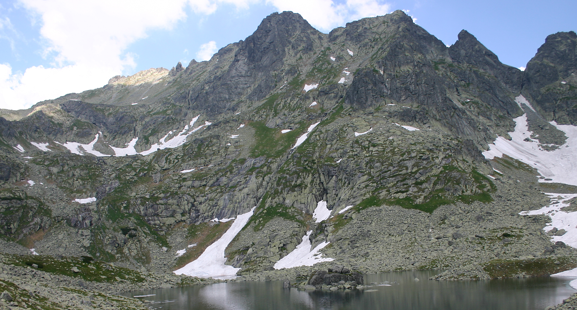 Fotografia ukazująca panoramę wysokich szczytów Tatr nad jeziorem Morskie Oko. Widoczne cztery szczyty górskie, z których najwyższy po lewej stronie, to Rysy. Stoki szczytów gdzieniegdzie są pokryte śniegiem.