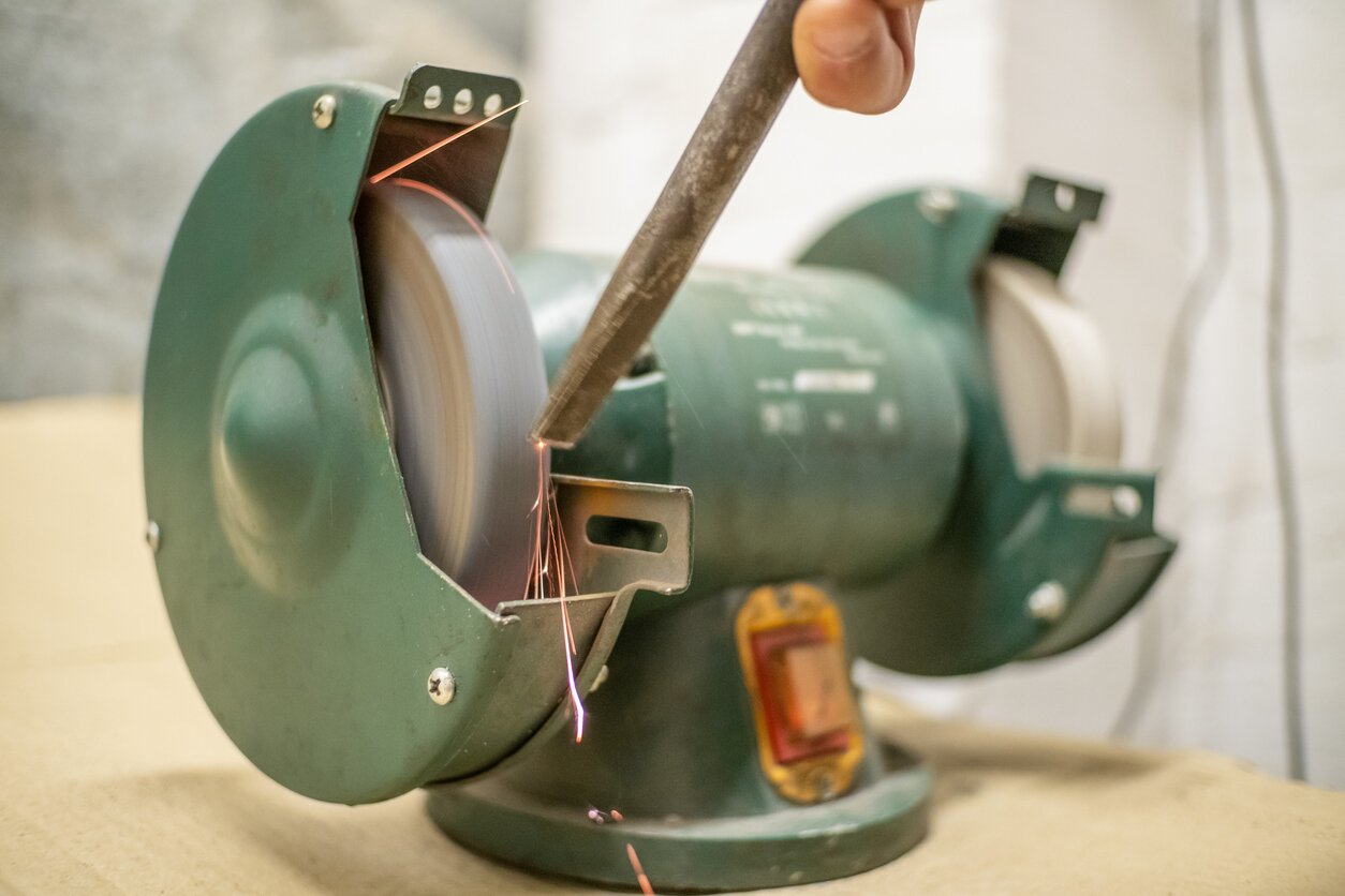 Zdjęcie przedstawia zieloną szlifierkę stacjonarną przymocowaną do stabilnego podłoża. Postać niewidoczna na ilustracji przykłada do ostrza szlifierki metalowy pręt, który poddaje obróbce.