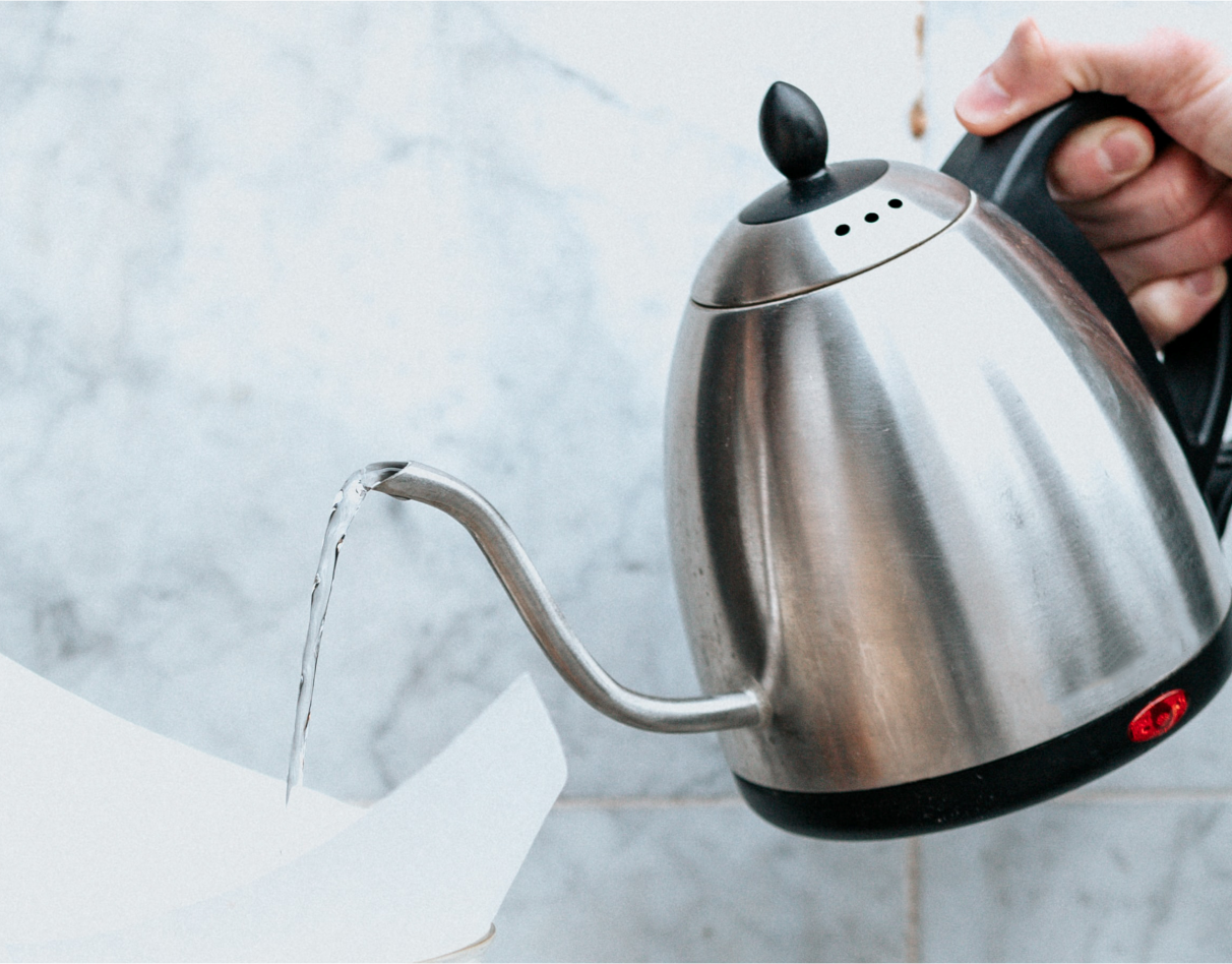 Dłoń trzymająca metalowy czajnik elektryczny w trakcie lania wody na filtr papierowy.