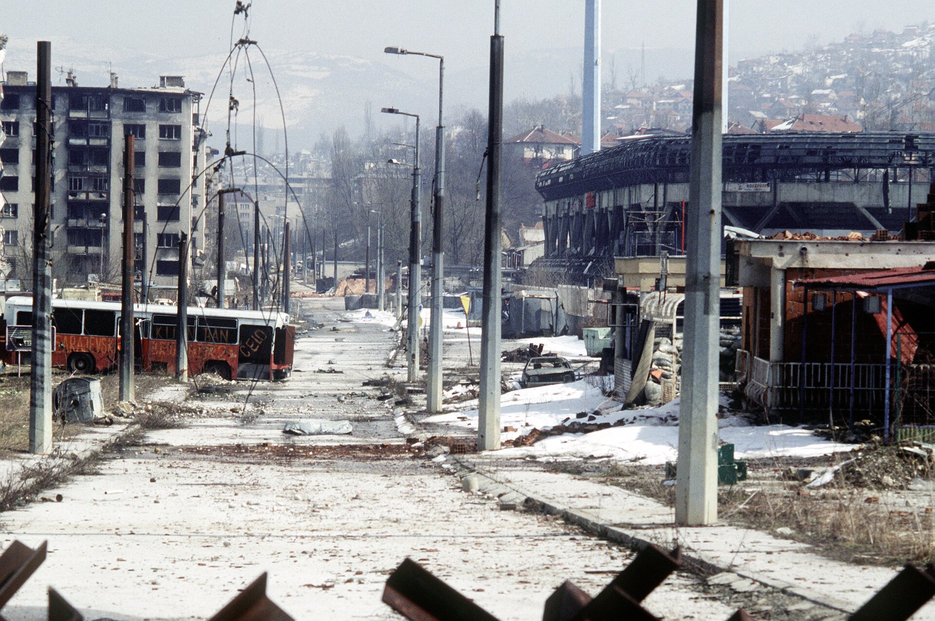 Zdjęcie przedstawia apokaliptyczny widok Sarajewa po wojnie w latach 1992 – 1996. W środku pusta pełna gruzów ulica, po bokach opalone latarnie, zniszczone i spalone szkielety bloków, sklepów, pojazdów. W okolicy nie żadnej osoby. Zdjęcie zrobione zza stalowej barykady.