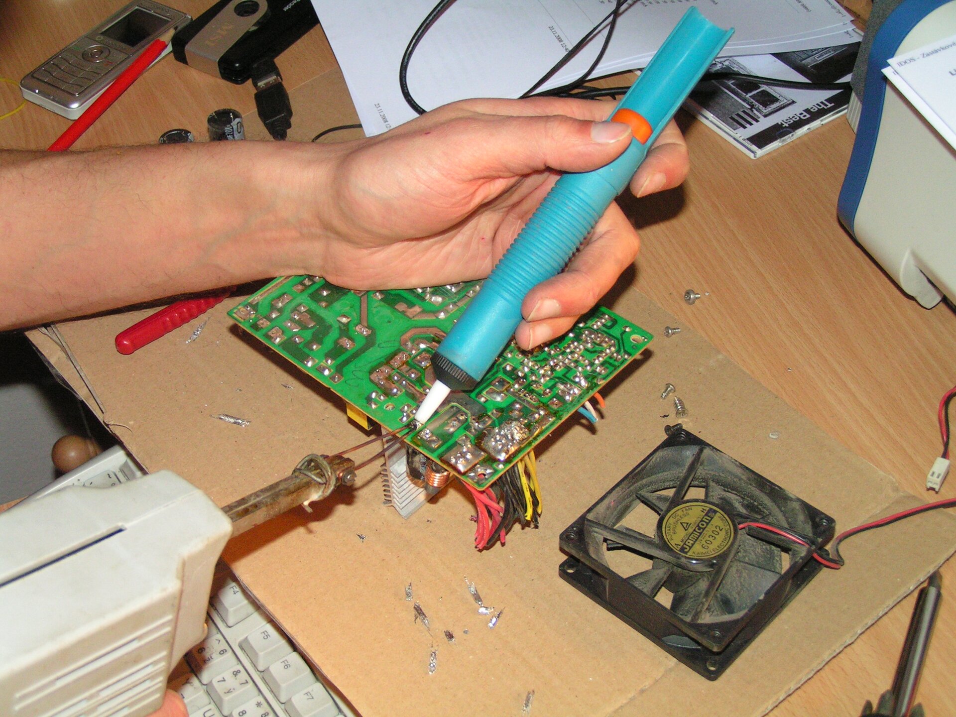 Zdjęcie przedstawia lutownicę pistoletową wraz z odsysaczem w rękach osoby odlutowującej komponenty z płytki PCB.