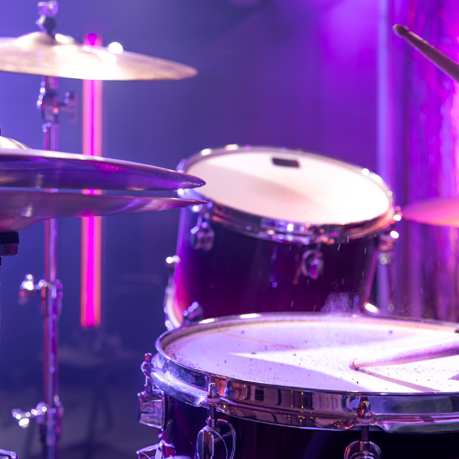 Ilustracja przedstawia zbliżenie na perkusję, w blasku ultrafioletu. Widoczne są talerze perkusyjne oraz bębny, w które uderzają pałeczki. 