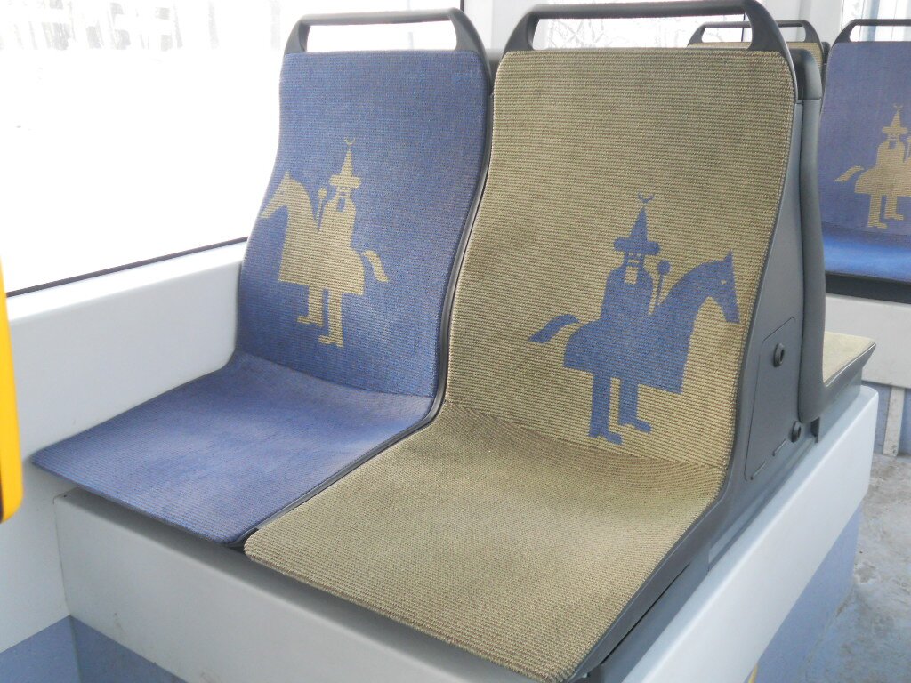 Kliknij, aby powiększyć. Kolorowe zdjęcie przedstawia wnętrze autobusu miejskiego. Widać dwa siedzenia, na oparciach których znajdują się podobizny lajkonika.