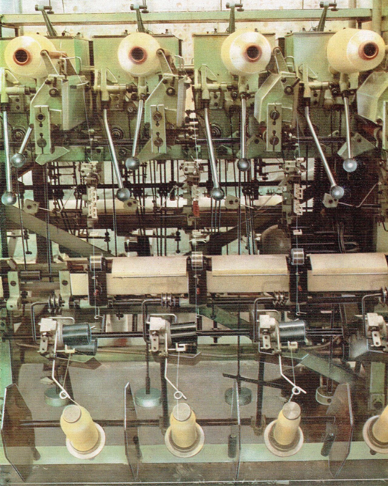 Zdjęcie przedstawia przewijarkę precyzyjną. Maszyna składa się z licznych, takich samych stanowisk przewijania. Na każdym stanowisku zamontowana jest szpulka nici w górnej części oraz szpulka nici w dolnej części. Przez wałki i inne elementy maszyny przechodzą nitki.