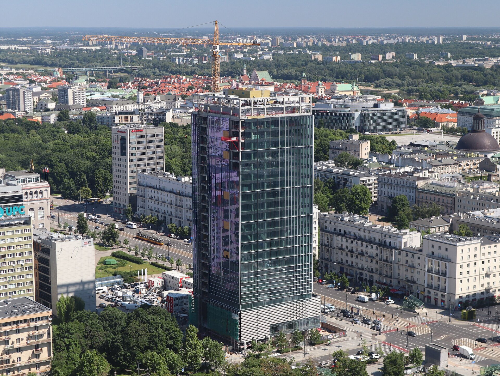 Zdjęcie przedstawia panoramę miasta. Na pierwszym planie wysoki szklany budynek. Obok niego znajduje się szeroki pas ulicy. Za nim wybudowane są bloki i niższe budynki kamienic.