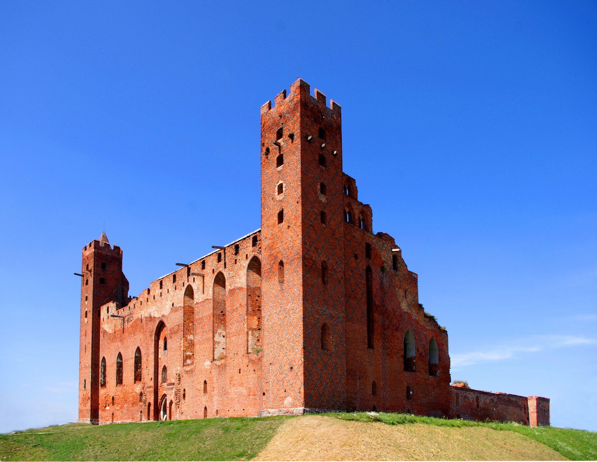 Zdjęcie przedstawia monumentalne ruiny zamku krzyżackiego na naturalnym wzniesieniu porośniętym trawą. Budowla zbudowana jest z czerwonej cegły na planie kwadratu z czworobocznymi wieżami w dwóch rogach zamku. Zdjęcie przedstawia ścianę zamku, pomiędzy wieżami, w której znajduje się brama wjazdowa. 
