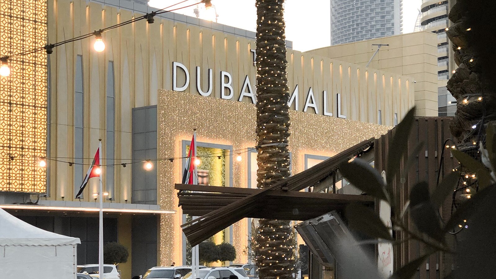 Zdjęcie przedstawia rozłożysty budynek, na elewacji którego widoczne są świecące się girlandy lampek. Pośrodku widoczne są wysokie wejścia, nad którymi widoczny jest napis DUBAI MALL. Przed budynkiem widoczne są pnie palm ozdobione światełkami. 