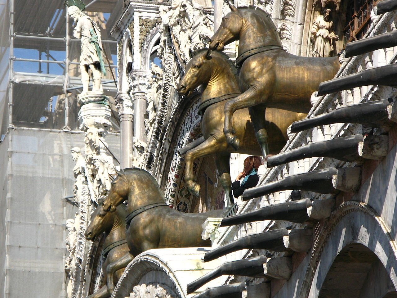 Ilustracja przedstawia rzeźby czterech, złotych koni w galopie, umieszczonych na fasadzie budynku. 