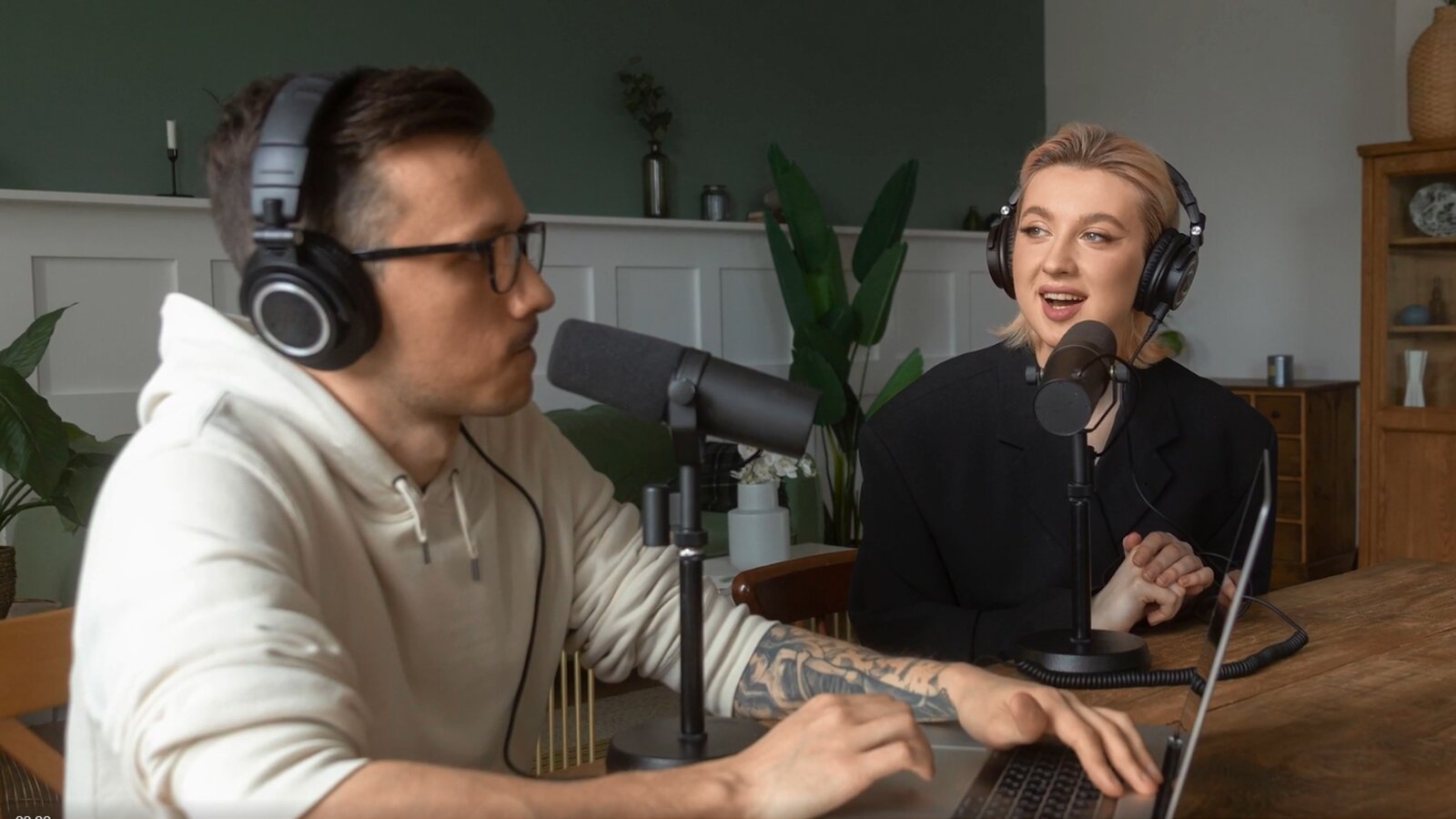 Zdjęcie przedstawia kobietę i mężczyznę siedzących przed mikrofonami przy stole, mających na uszach profesjonalne słuchawki.