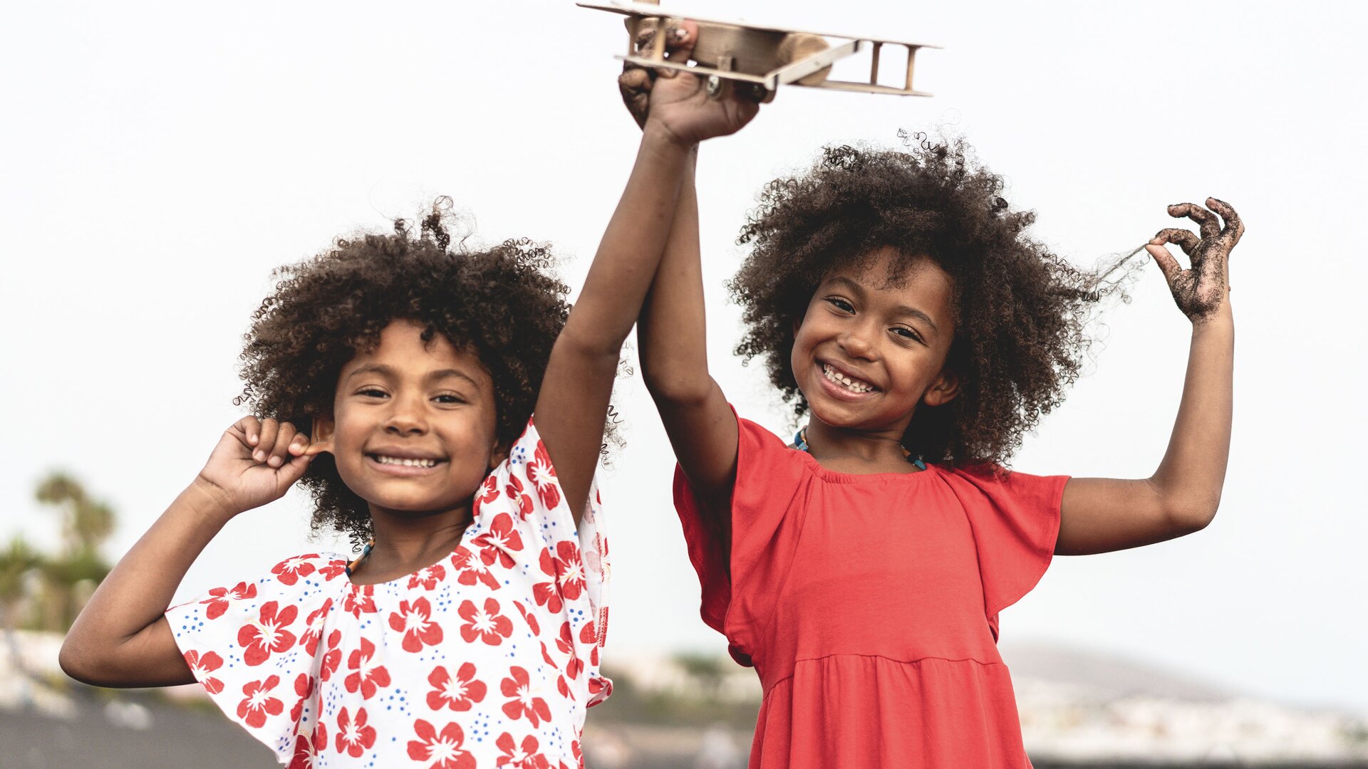 Zdjęcie przedstawia bliźniaczki, które bawią się drewnianym samolocikiem. Dziewczynki mają ciemne, kręcone włosy.