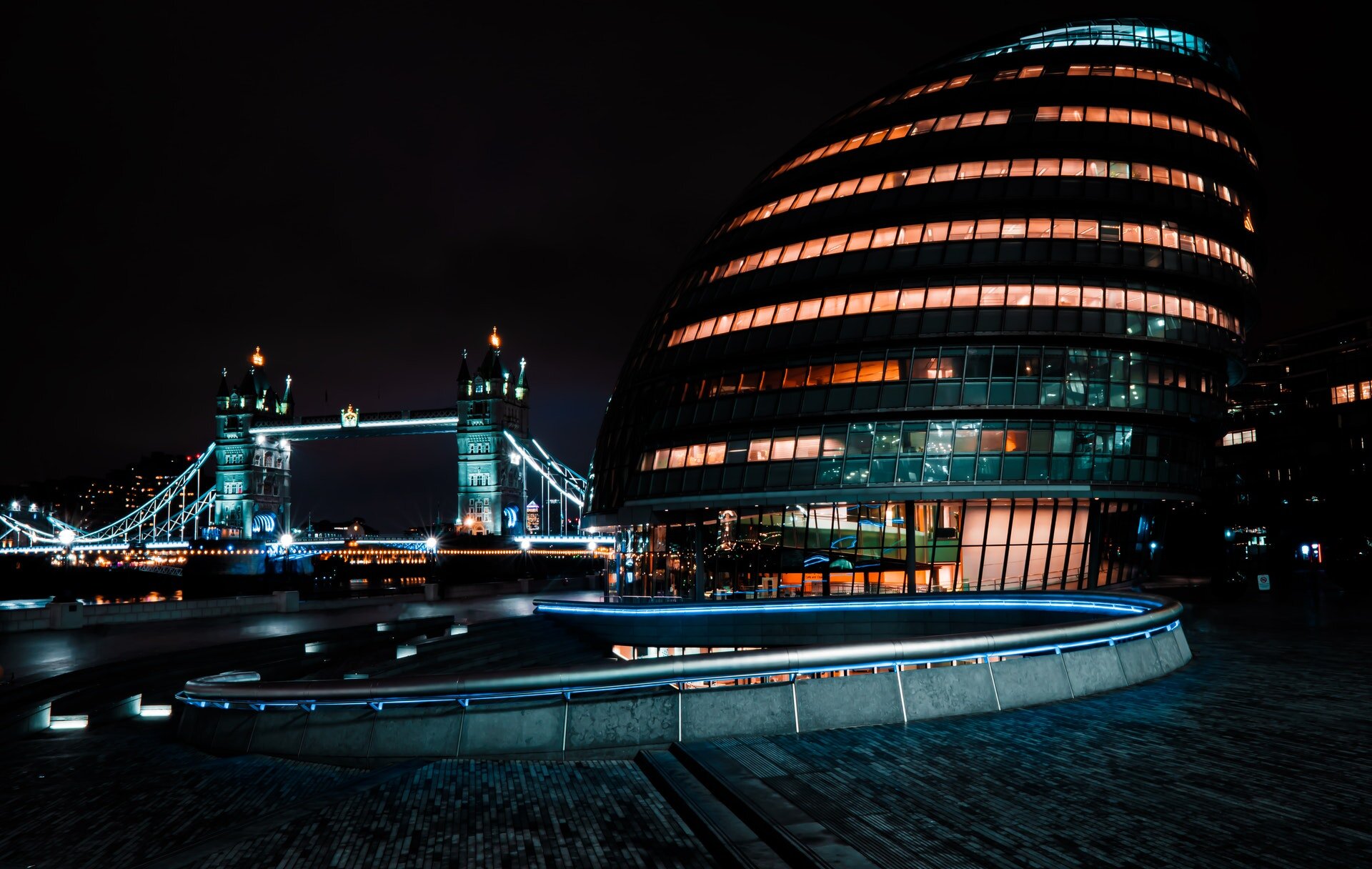 Fotografia przedstawia Londyn nocą. Na pierwszym planie po prawej stronie zdjęcia znajduje się City Hall. Budynek ma owalny, jajowaty kształt. Posiada kilka pięter, na każdym z nich widnieje szereg oświetlonych okien. W tle znajduje się wysoki, oświetlony most z dwoma wieżami połączonymi ze sobą. 