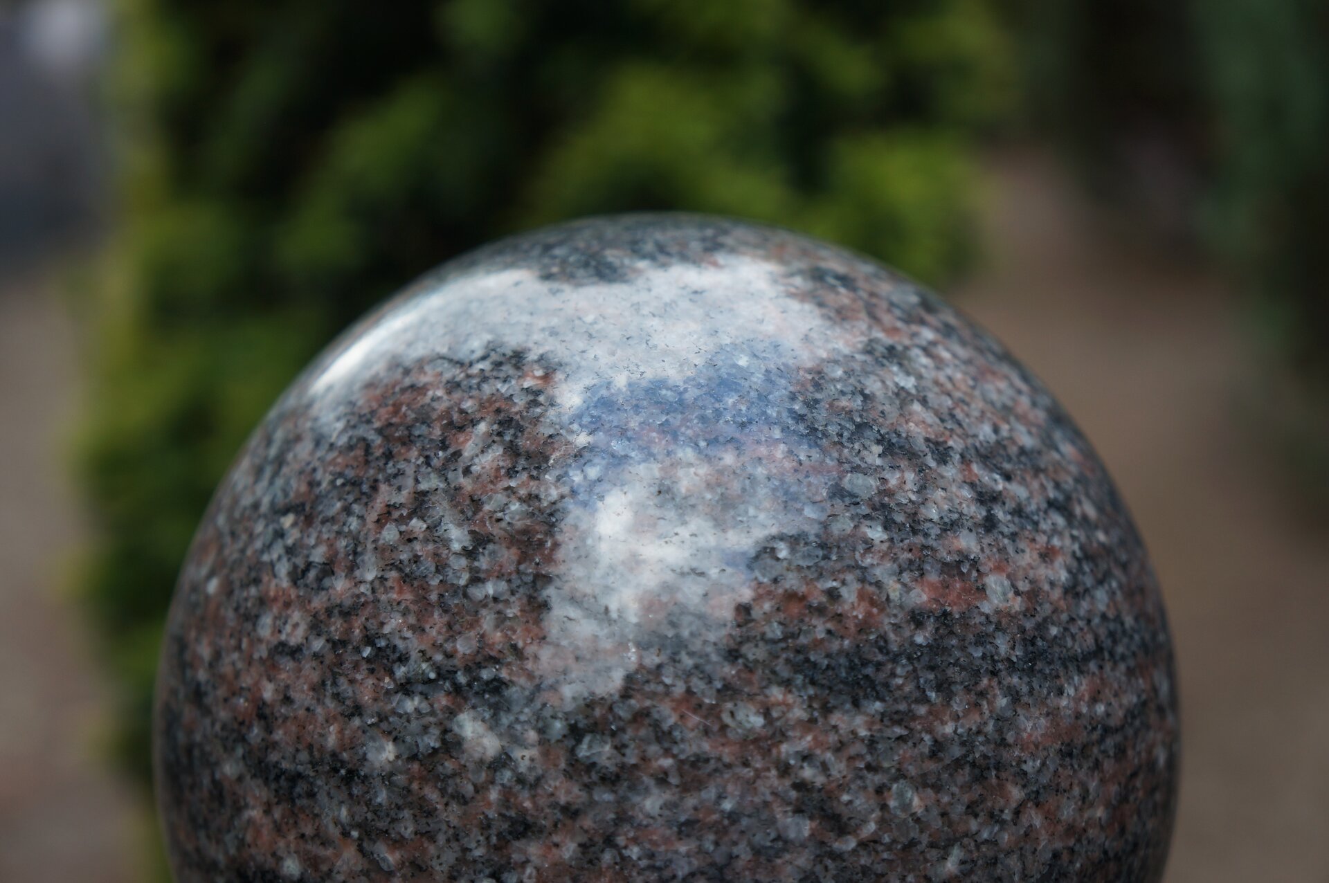 Ilustracja przedstawia rzeźbę pt. „Kula”. Na zdjęciu widoczny jest fragment kuli wykonanej z granitu. Na brązowo-biało-czarnej powierzchni odbija się światło. Tło jest rozmazane.