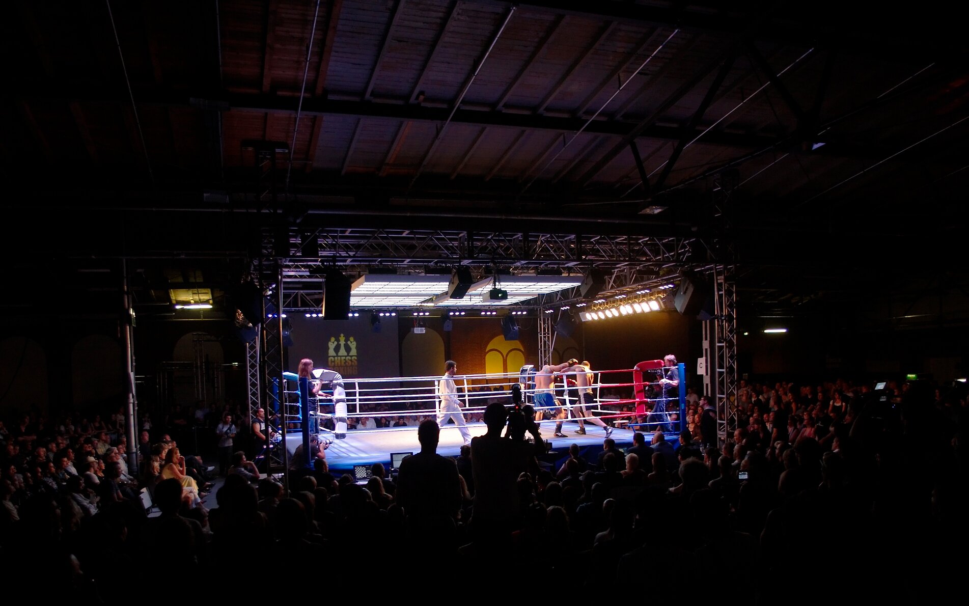 Zdjęcie przedstawia oświetlony ring, na którym widoczni są dwaj walczący bokserzy. Obok nich stoi mężczyzna ubrany w biały strój i obserwuje walkę. Wokół ringu widoczni są kibice obserwujący walkę. Na ścianie widoczny jest znak pionka szachowego i napis w języku angielskim: CHESS .