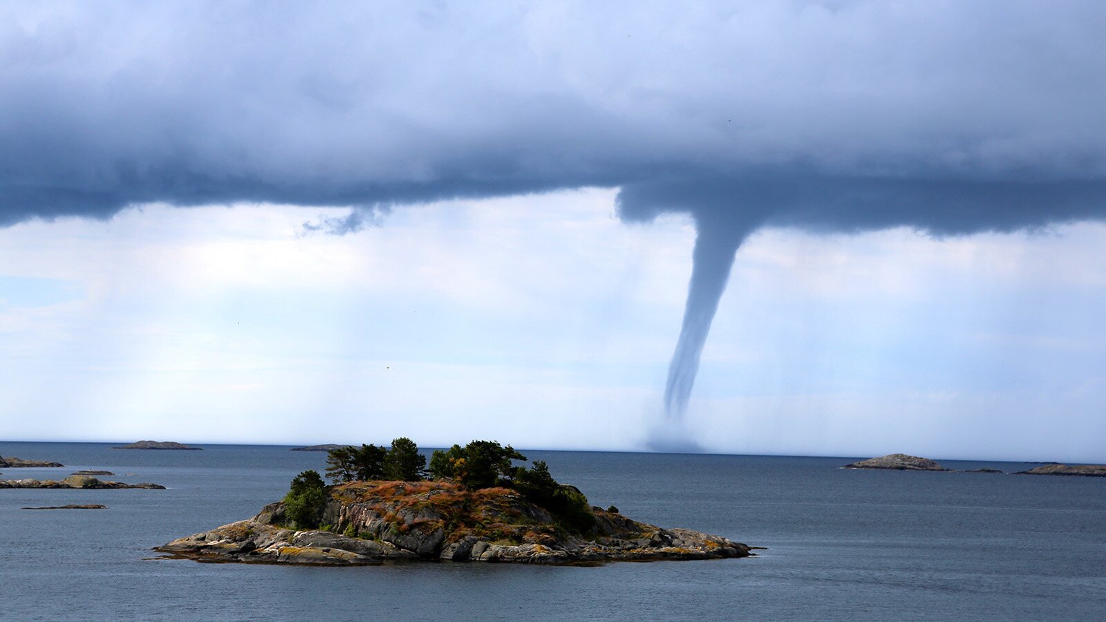 Zdjęcie przedstawia zbiornik wodny z niewielką, skalistą wysepką na pierwszym planie i pojedynczymi skałami widocznymi w tle. Woda ma kolor ciemnogranatowy. Na horyzoncie widoczne jest zasnute fioletowymi chmurami niebo oraz trąba powietrzna, która utworzyła się nad taflą wody. W miejscu styku trąby powietrznej z wodą widoczne jest kłębowisko fioletowej wody.