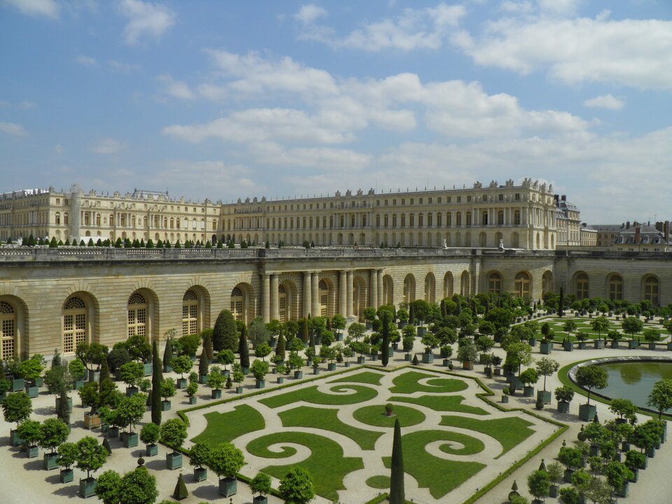 Widok na ogrody Wersalu z geometrycznym parterem z niskich żywopłotów i drzewami w donicach ustawionymi w równych rzędach. W tle widać długą fasadę pałacu oraz arkadową galerię oranżerii. Nad ogrodami rozciąga się jasne niebo z rozproszonymi chmurami.