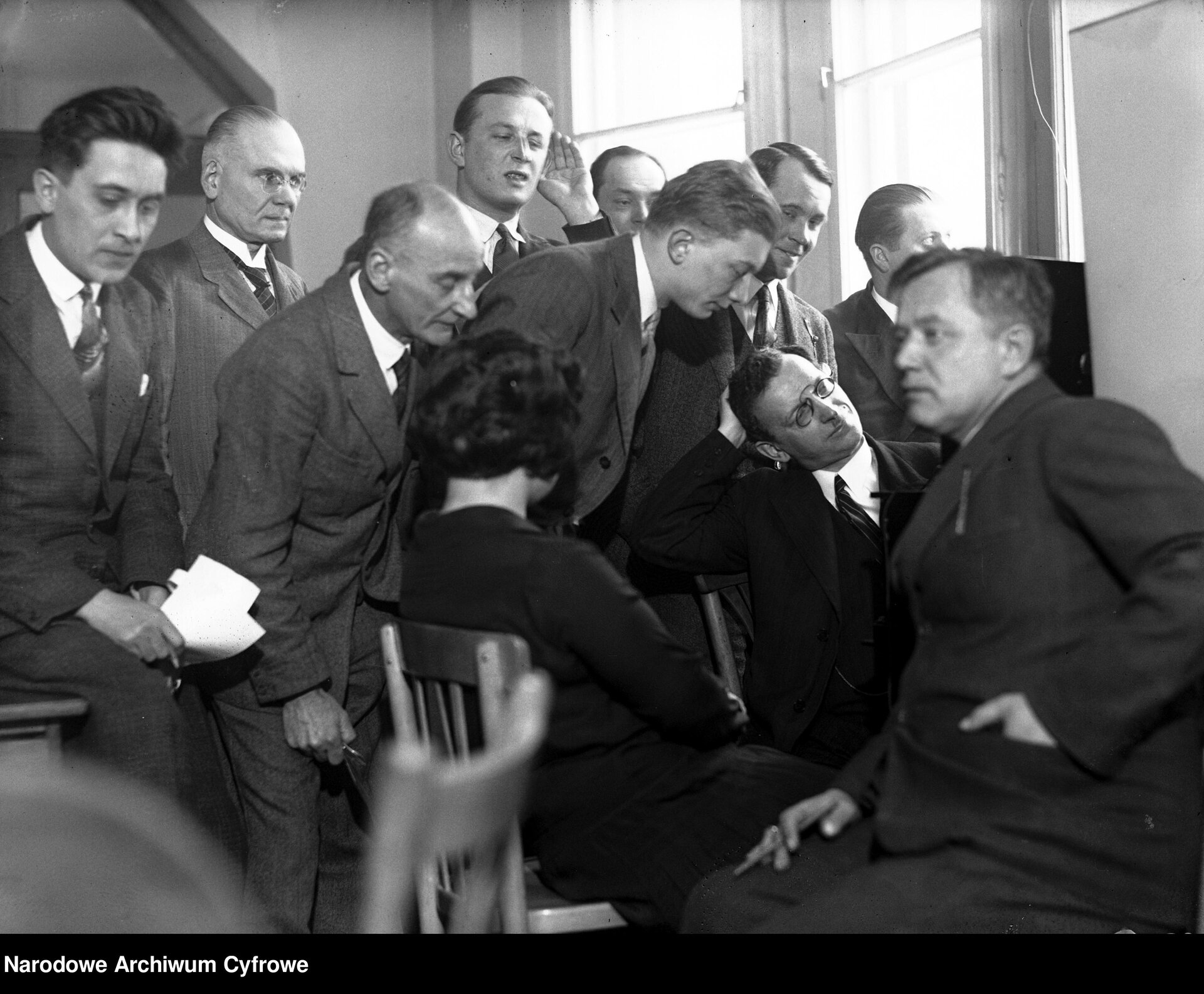 Zdjęcie przedstawia grupę mężczyzn w garniturach i jedną kobietę, którzy znajdują się w pokoju. Siedzą i stoją przy radiu. Słuchają.