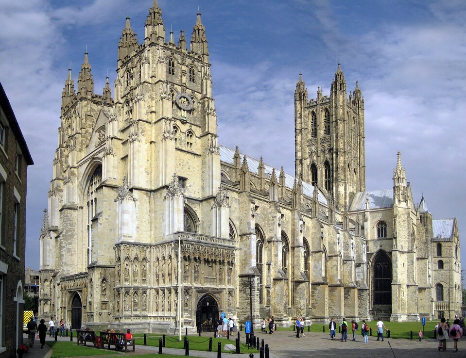 Ilustracja o kształcie poziomego prostokąta przedstawia bryłę katedry Canterbury. Wykonane od strony zachodnio‑południowej zdjęcie ukazuje portal wejściowy i fasadę od południa z widocznym transeptem. Na skrzyżowaniu naw z transeptem znajduje się wieża, a jej naroża dekorowane są wieżyczkami, podobnie jak wieże portalowe. Przypory odpowiadają podziałowi wewnętrznemu na przęsła. Okna zakończone są łukiem ostrym. Przed kościołem spacerują ludzie.