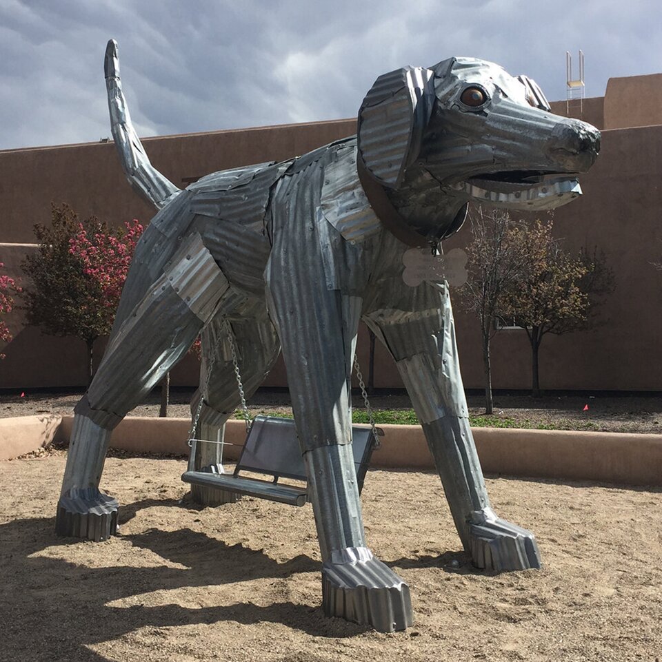 Fotografia przedstawia rzeźbę - psa, na tle kremowego muru oraz niewielkich drzew z różowymi kwiatami. rzeźba jest ponadnaturalnej wysokości i sięga pierwszego piętra - łeb psa jest na równi z dachem budynku, obok którego stoi. Pies stoi w ogrodzonym fragmencie przestrzeni, które wysypane zostało piaskiem. Rzeźbę wykonano z blachy falistej o nieregularnej strukturze, na której można dostrzec wiele linii oraz wgłębień. Do brzucha psa łańcuchem przytwierdzono huśtawkę. Pies ma brązowe oczy oraz obrożę z zawieszoną na niej kością.