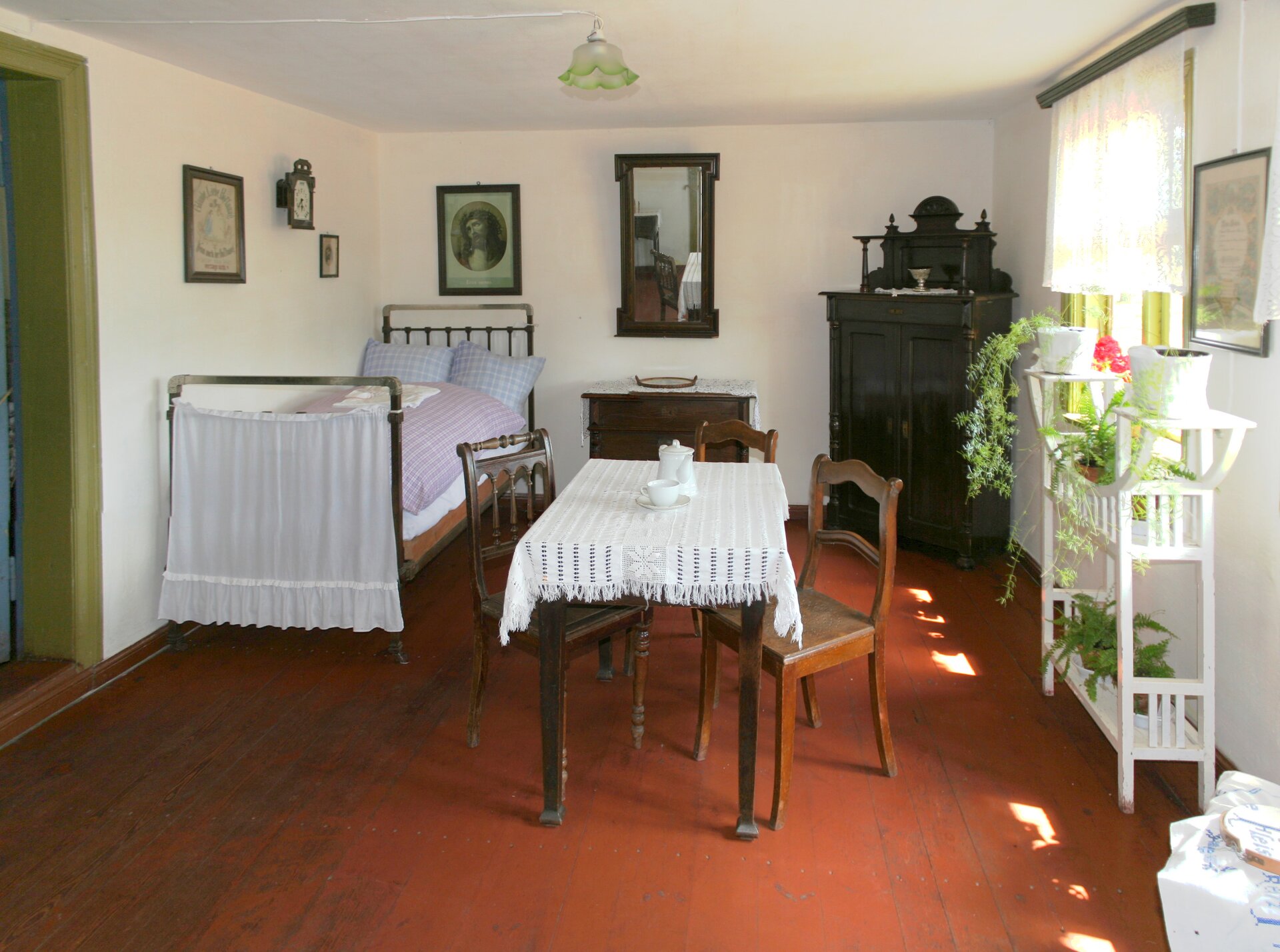 Zdjęcie przedstawia izbę mieszkalną z Muzeum Wsi Słowińskiej w Klukach. W pomieszczeniu na środku ukazany jest stój nakryty obrusem, przy którym stoją trzy drewniane krzesła. Po lewej stronie pomieszczenia znajduję się jednoosobowe zasłane łóżko. Zaraz obok stoi komoda, nad którą wisi lustro. W lewej części pokoju w rogu, z ukosa widnieje drewniana szafa. Na lewej ścianie znajduję się okno, przy którym stoi stojak z kwiatami. Na ścianach wiszą obrazy a ze środka sufitu zwisa mała zielona lampka.