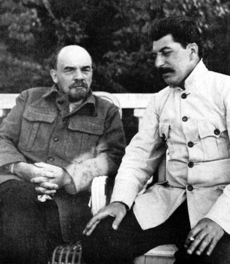 Zdjęcie przedstawia dwóch siedzących obok siebie mężczyzn na ławce. Są to Lenin po lewej i Stalin po prawej. Lenin jest starszy, łysiejący, ma brodę z wąsami, ubrany jest w płaszcz, ma złączone ręce. Stalin ma ciemne, zaczesane do tyłu włosy, gęste wąsy i jasny mundur, ma oparte ręce na nogach. Za mężczyznami widoczne są drzewa.