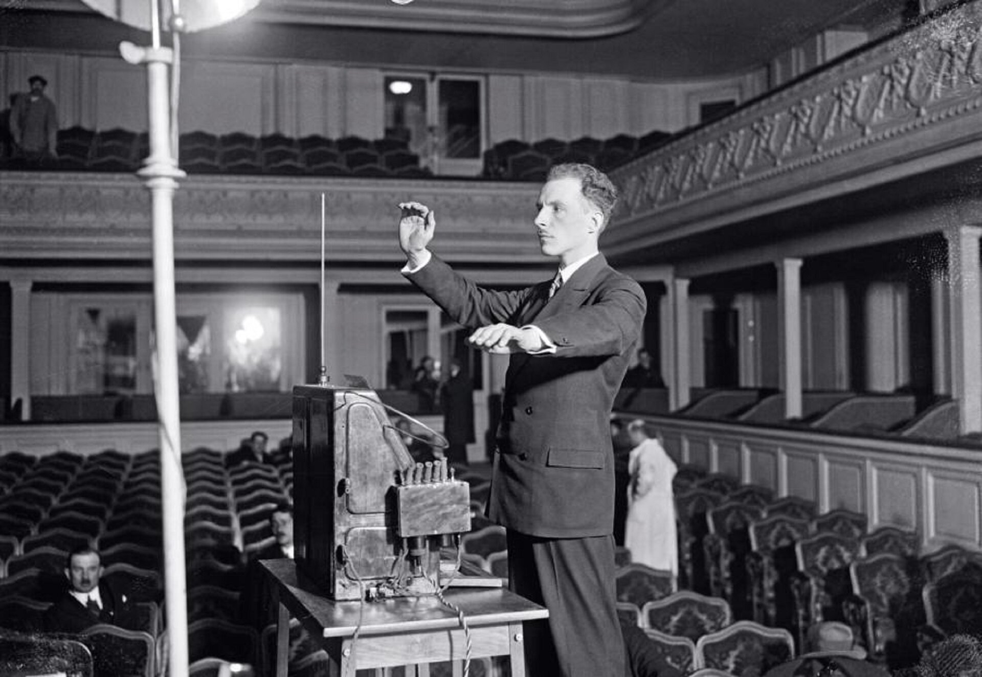 Ilustracja przedstawia rosyjskiego wynalazcę - Lew Siergiejewicz Termen. W 1927 roku zbudował najbardziej zaawansowany prototyp telewizora. Mężczyzna stoi na scenie przed instrumentem w pustej sali operowej. Ubrany jest elegancko. Ręce ma uniesione ku górze, gra na thereminie.