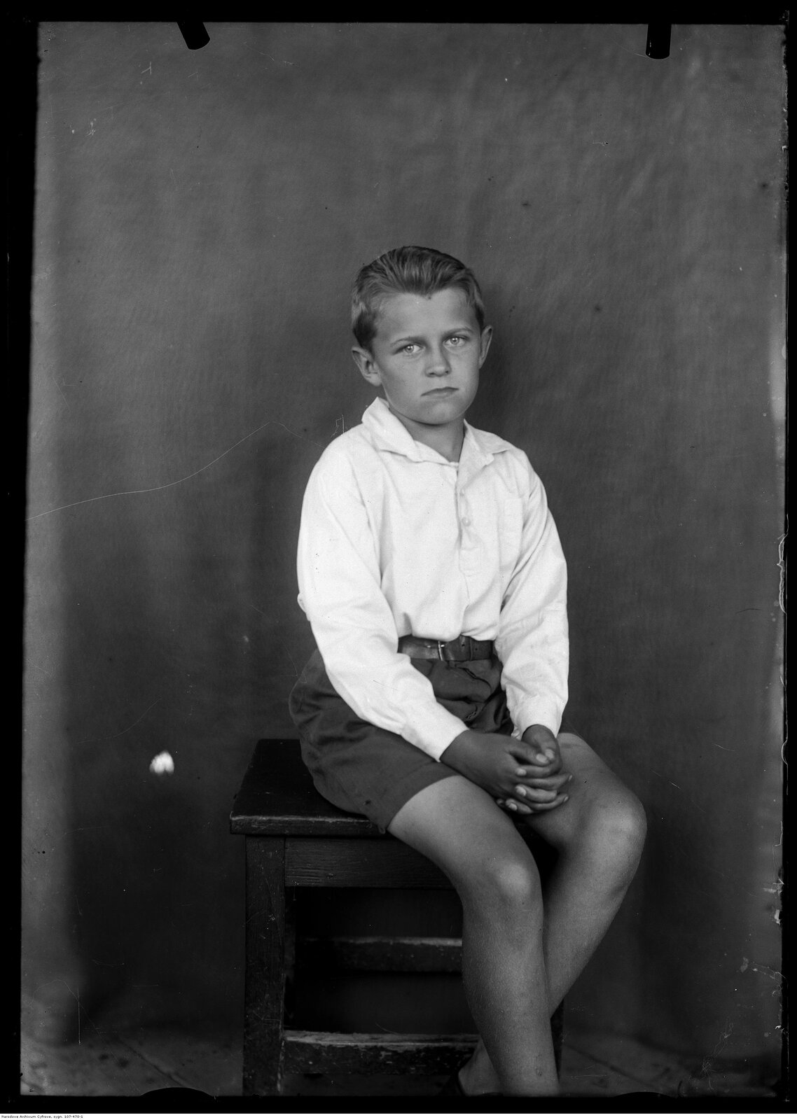 Na zdjęciu znajduje się siedzący na krześle jasnowłosy chłopiec w białej koszuli i krótkich spodenkach. Na drugim planie w tle rozwieszona tkanina.