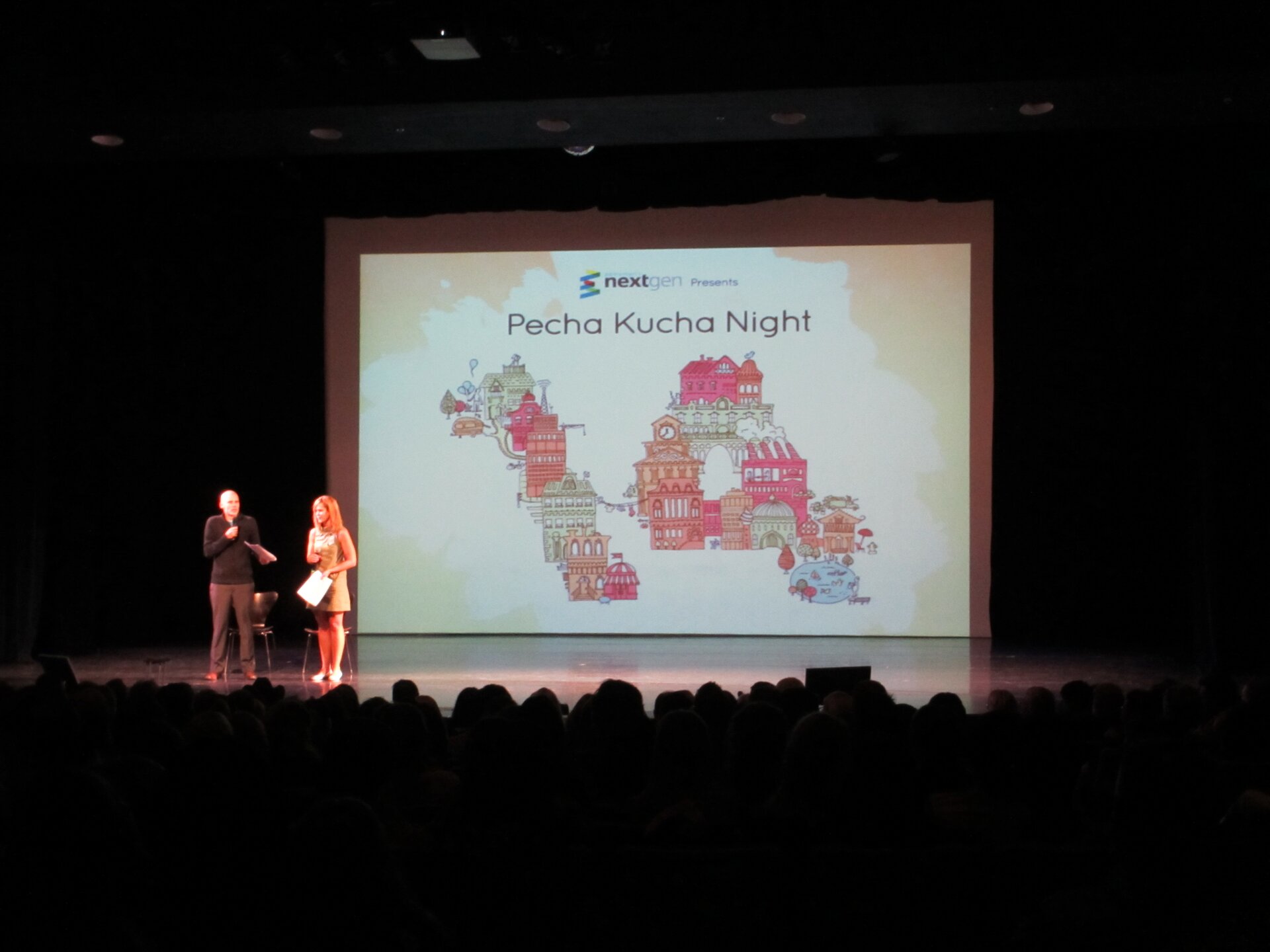 Zdjęcie przedstawia dwie osoby na scenie oglądane przez widownię. Za prezentującymi osobami wyświetla się prezentacja o tytule Pecha Kucha Night.