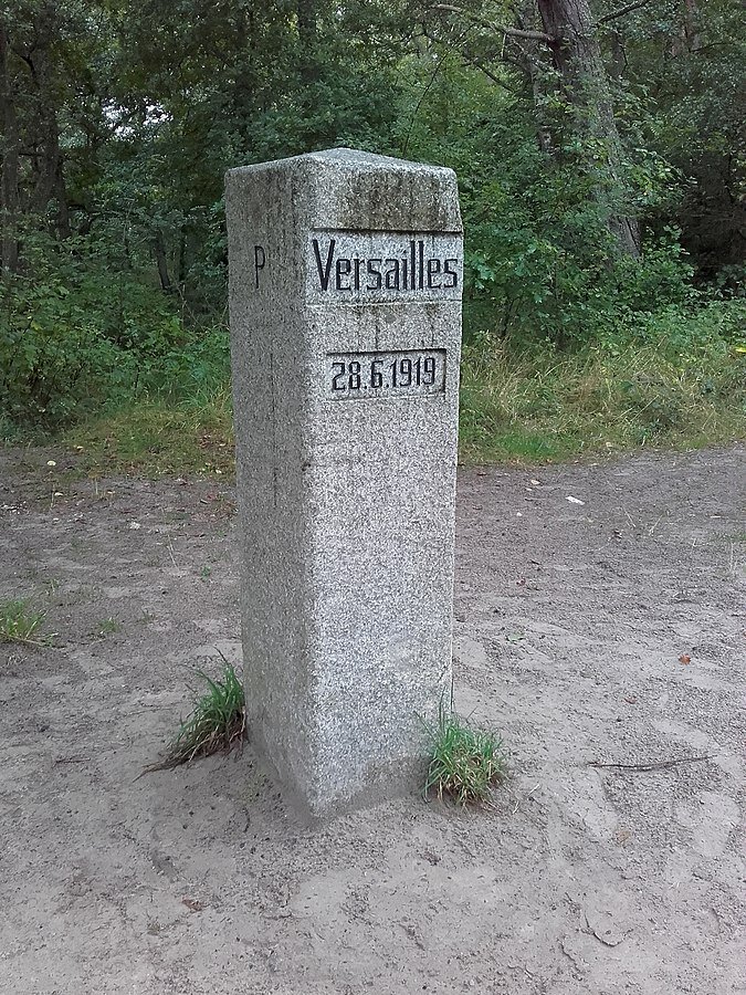 Zdjęcie przedstawia wmurowany w ziemię kamienny słup o kształcie prostopadłościanu. Widnieje na nim wyryty napis Versailles 28.6.1919. Za słupem widoczny jest las. 